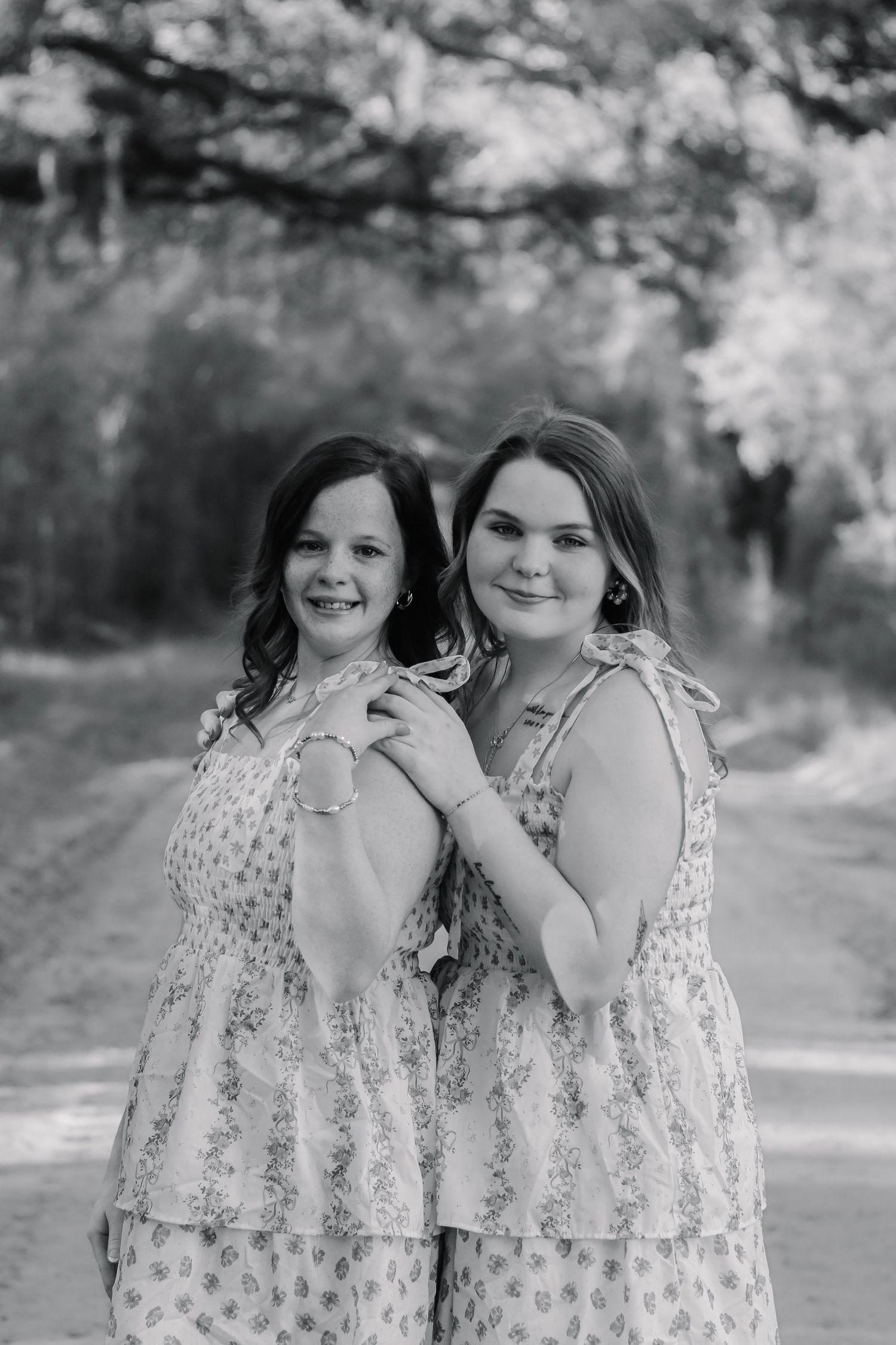 Black and white portrait of two smiling girls in matching floral dresses on a dirt road.