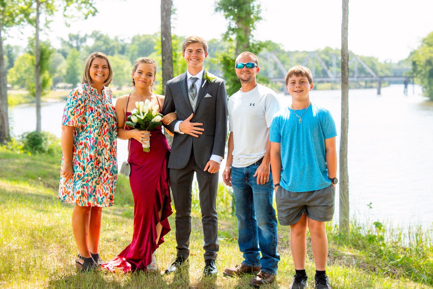Family of five poses outdoors near river with prom couple in formal red gown and gray suit.