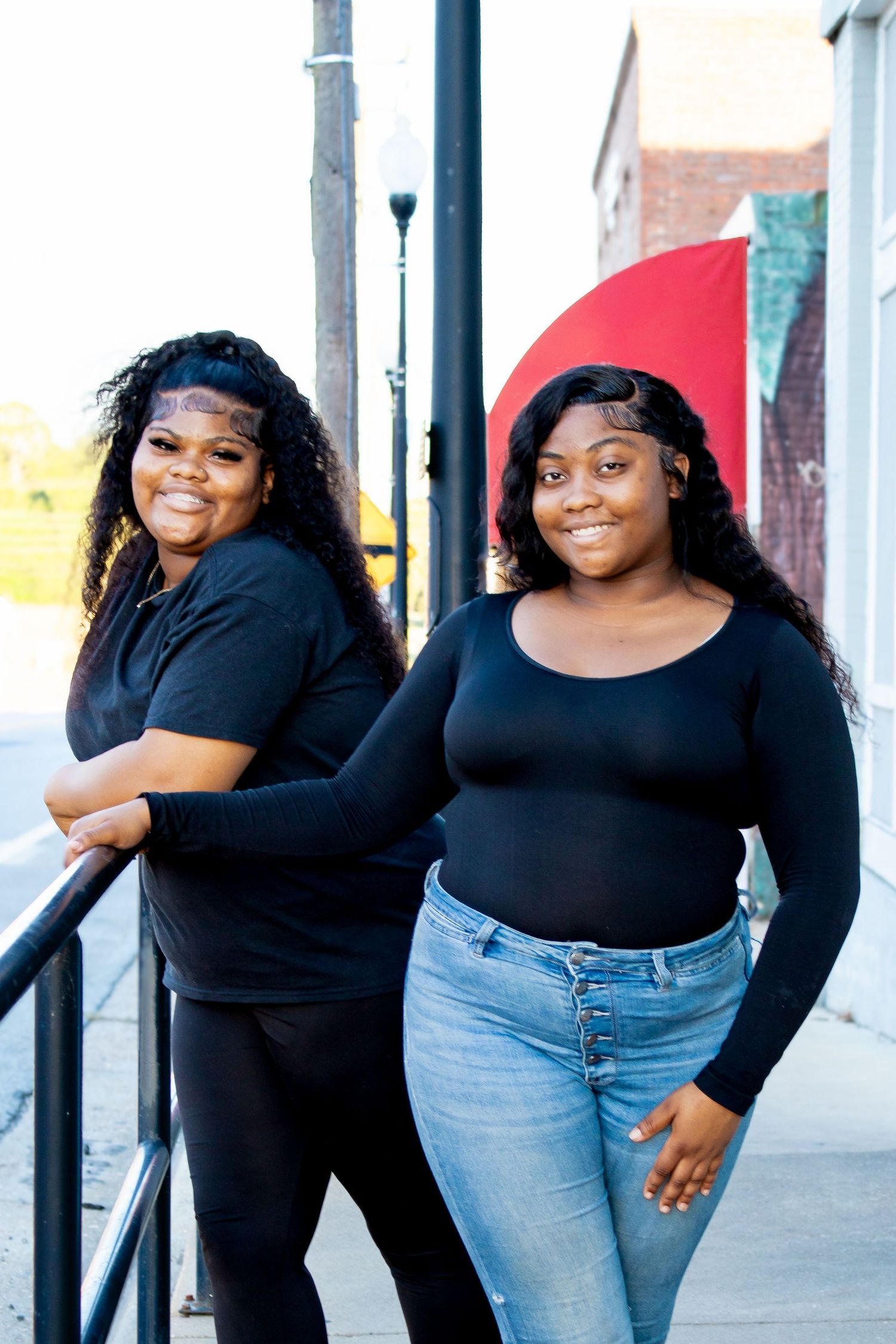 Two young Black women in black outfits posing outdoors on a sidewalk with a colorful mural behind them.