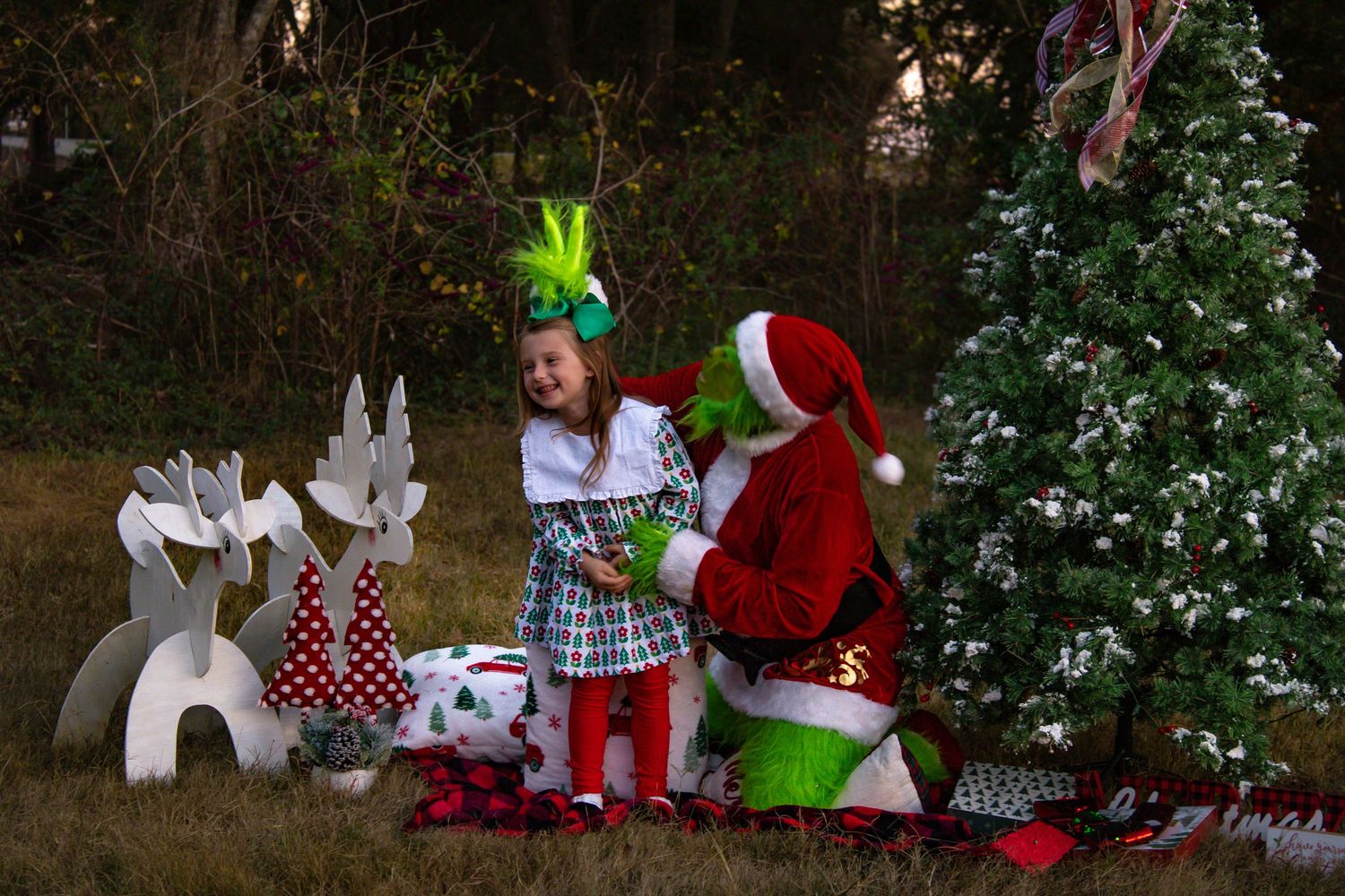 Young girl with green bow poses with Grinch character beside Christmas tree and reindeer props.