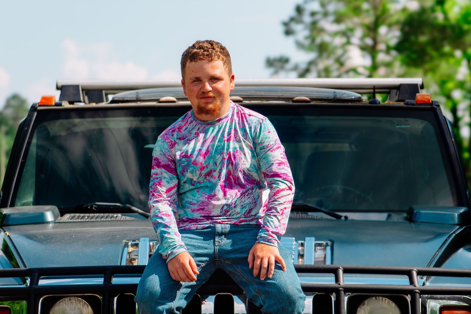 Man in vibrant tie-dye long sleeve shirt sitting on the hood of a dark green Hummer H2 outdoors.