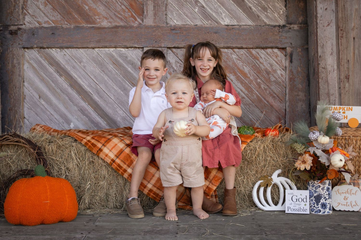 Four siblings posing together on hay bale with pumpkins in festive fall barn photo session.