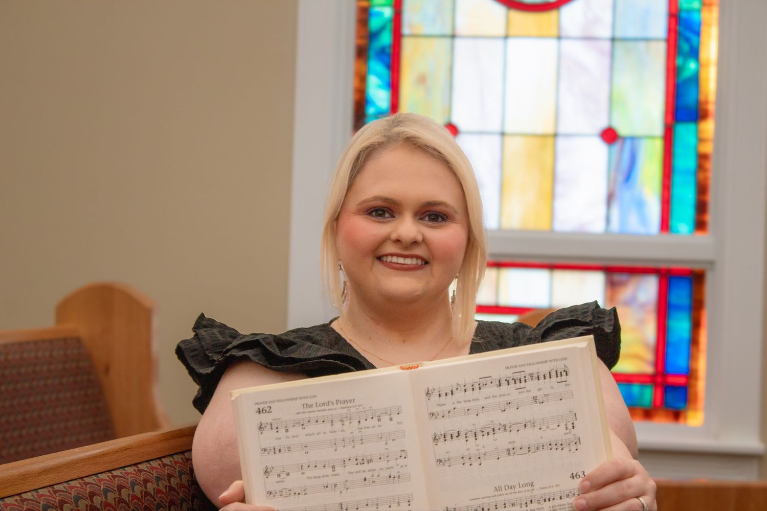 Young blonde woman smiling and holding open a sheet music book inside a church with stained glass windows.