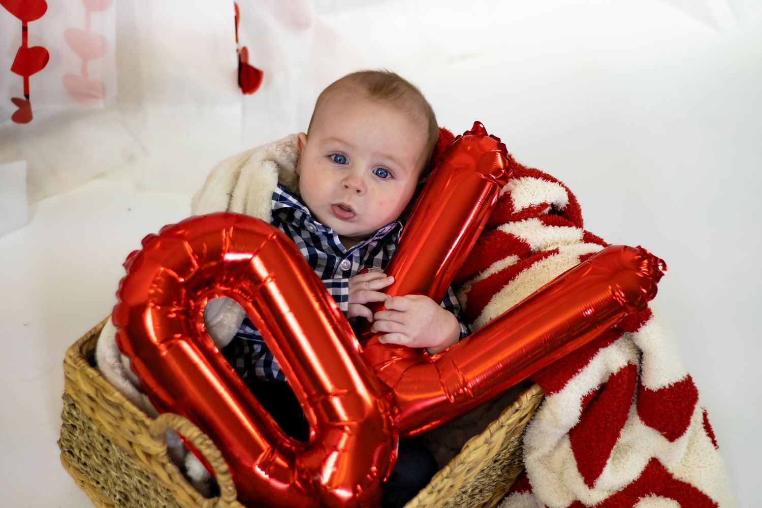 Cute baby sitting in a wicker basket holding red number balloon props for a Valentine's Day photo.