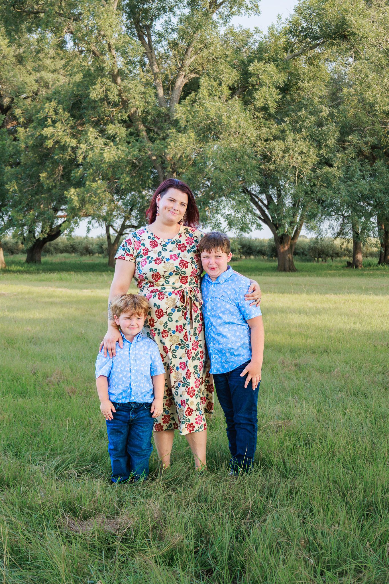 A mother in a floral dress poses outdoors with her two young sons in matching blue shirts on a green lawn.