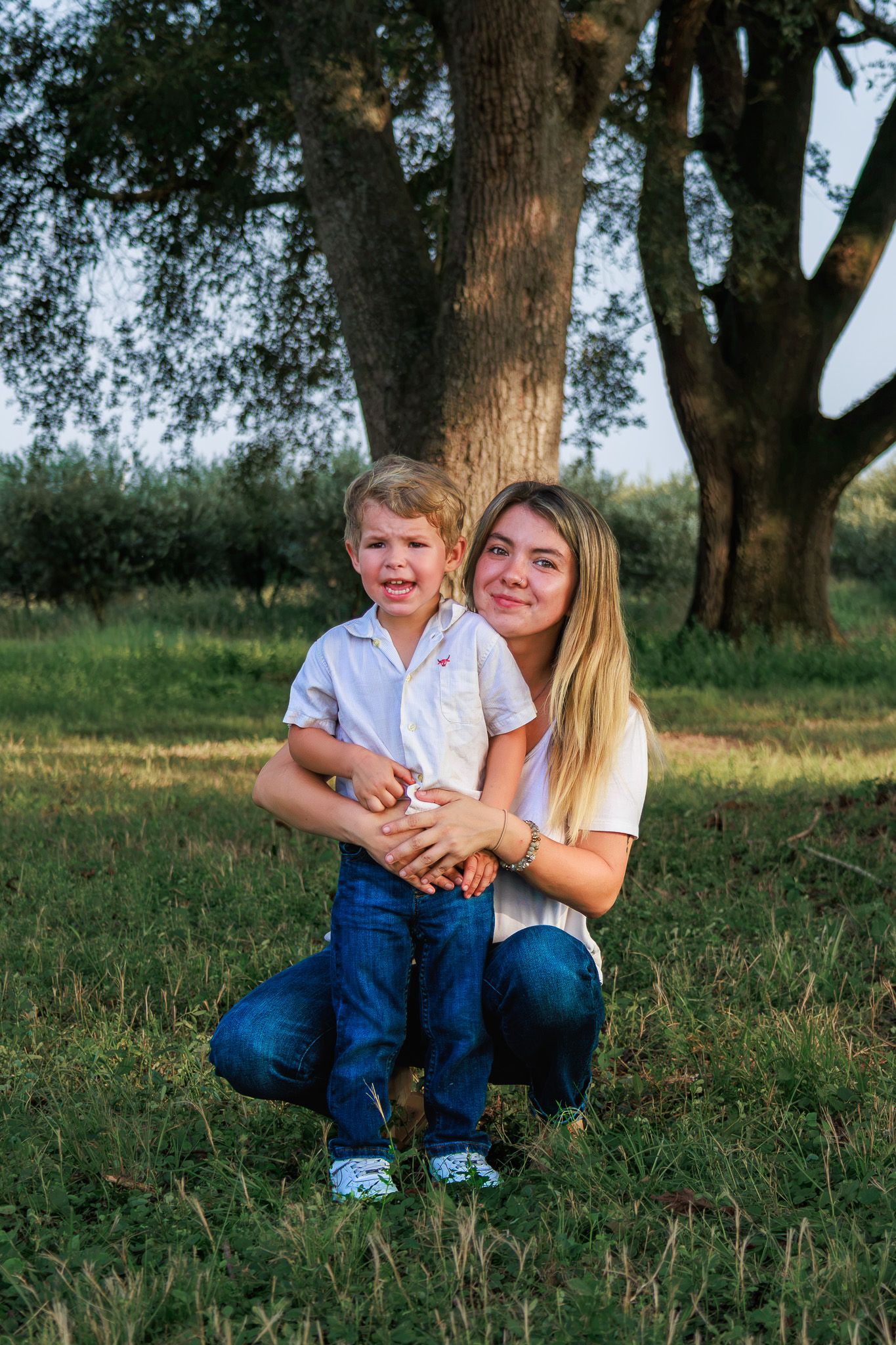 A young woman crouches behind a smiling toddler boy outdoors near a large tree on green grass.