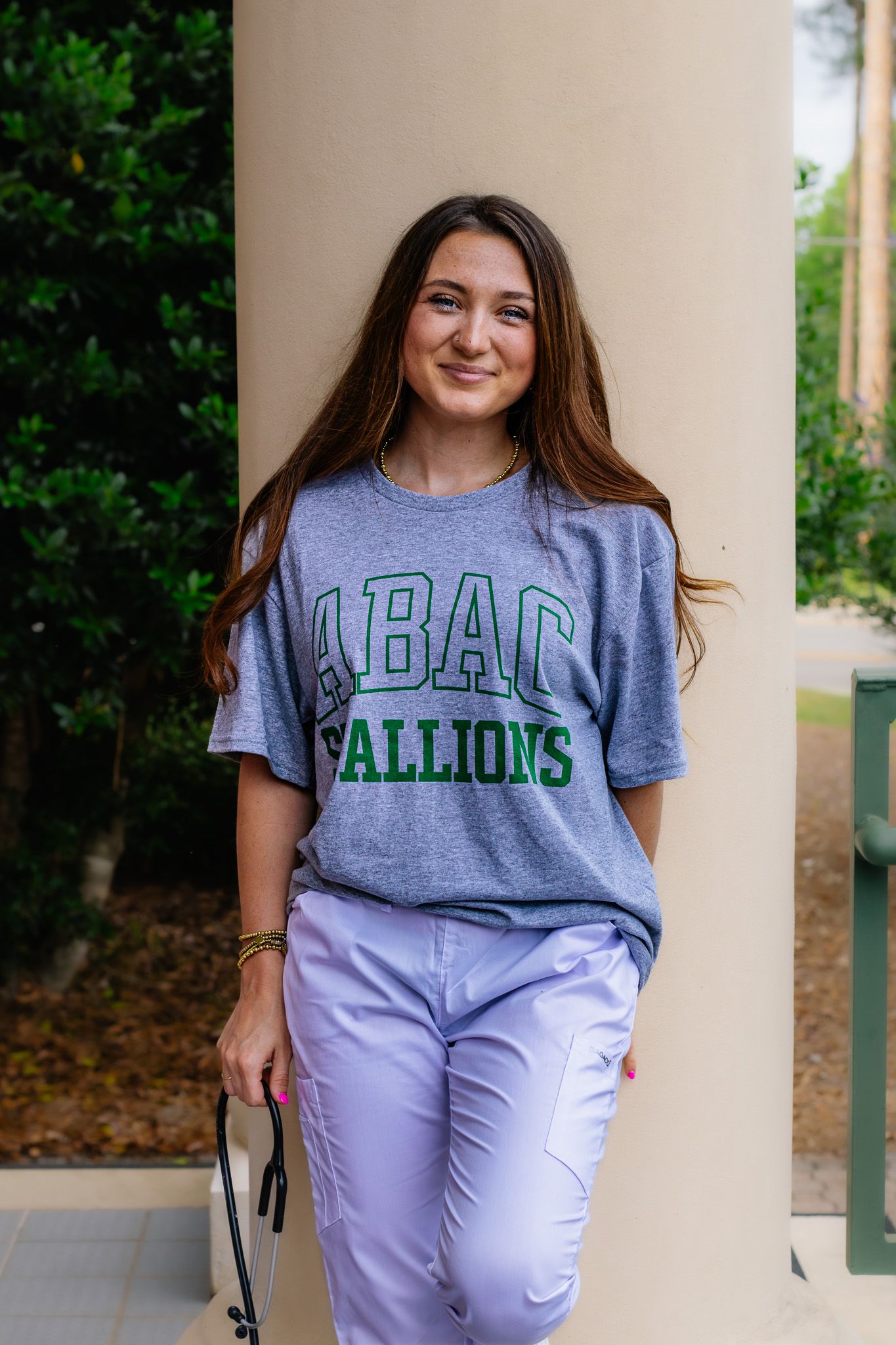 Smiling woman in ABAC Stallions gray t-shirt and lavender pants leans against white column outdoors.