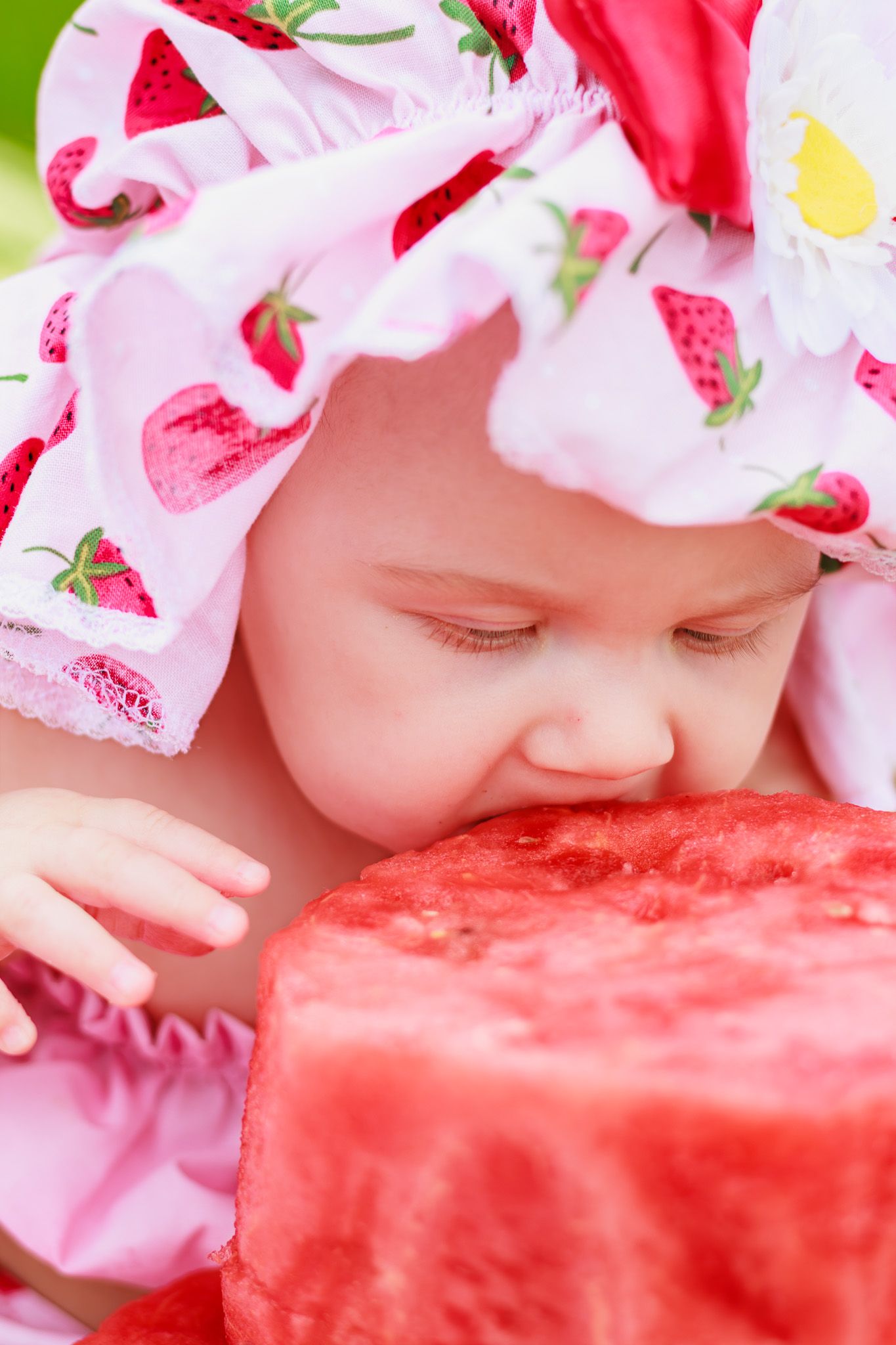 Cute baby in pink strawberry-print hat biting into a fresh slice of juicy red watermelon outdoors.
