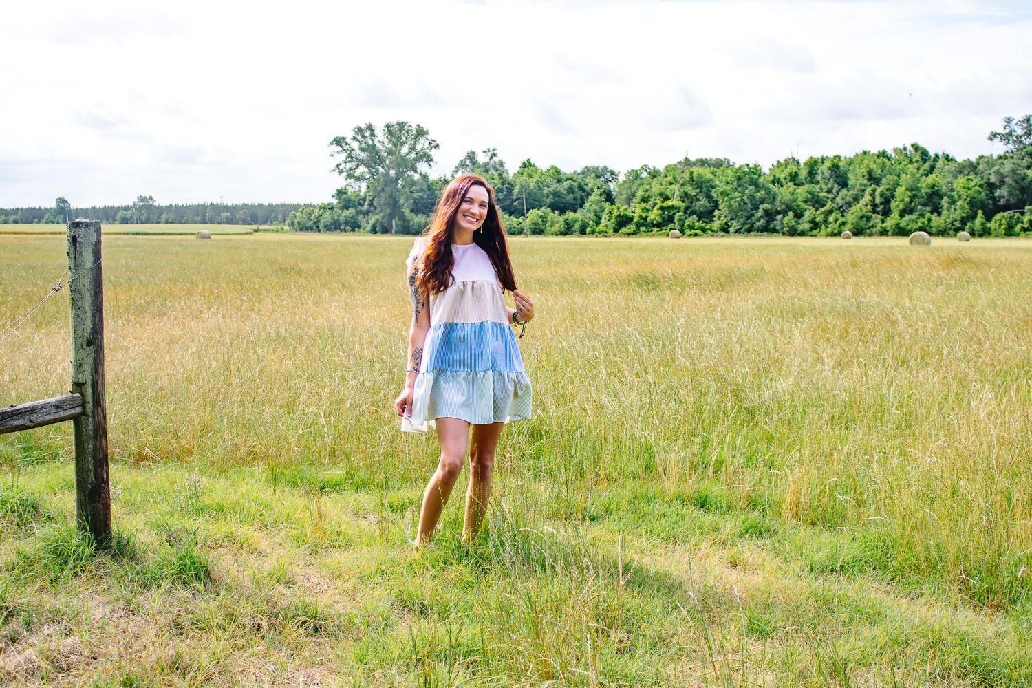 A smiling teenage girl in a colorful dress stands near a wooden fence post in a wide open grassy field.