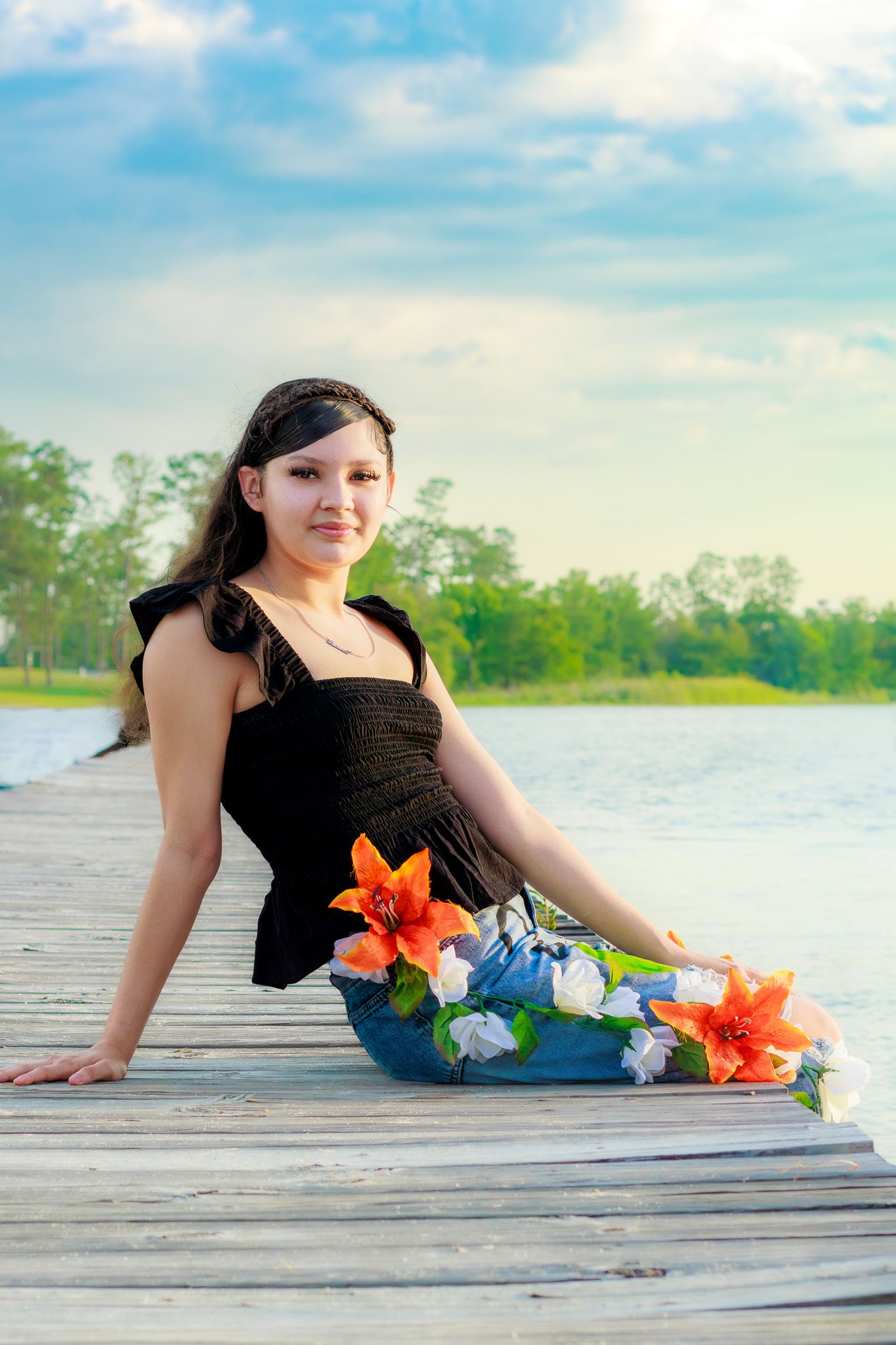 Young woman in black top and floral jeans sits on a wooden dock by a calm lake, holding orange lilies under a blue sky.