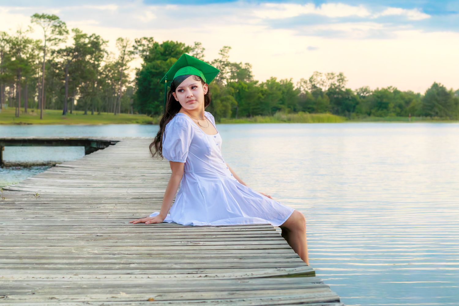 A young woman in a white dress and green graduation cap sits on a wooden dock by a serene lake at sunset.
