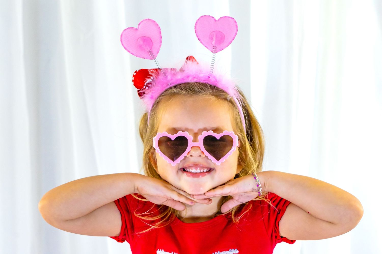 Cute girl in red shirt wearing pink heart sunglasses and heart headband for Valentine's Day portrait.