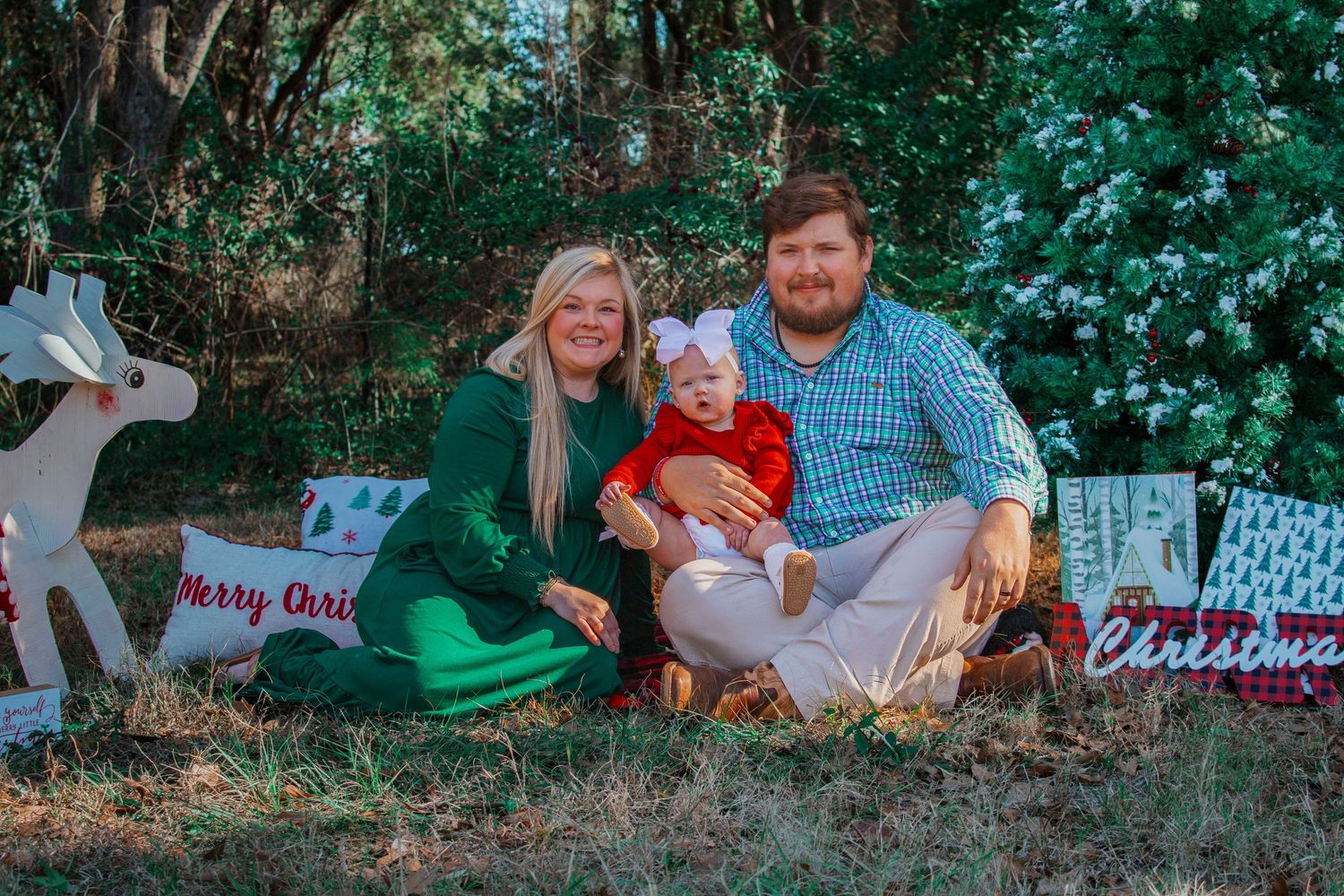 Family of three with baby girl in red dress posing with Christmas signs in an outdoor holiday portrait.