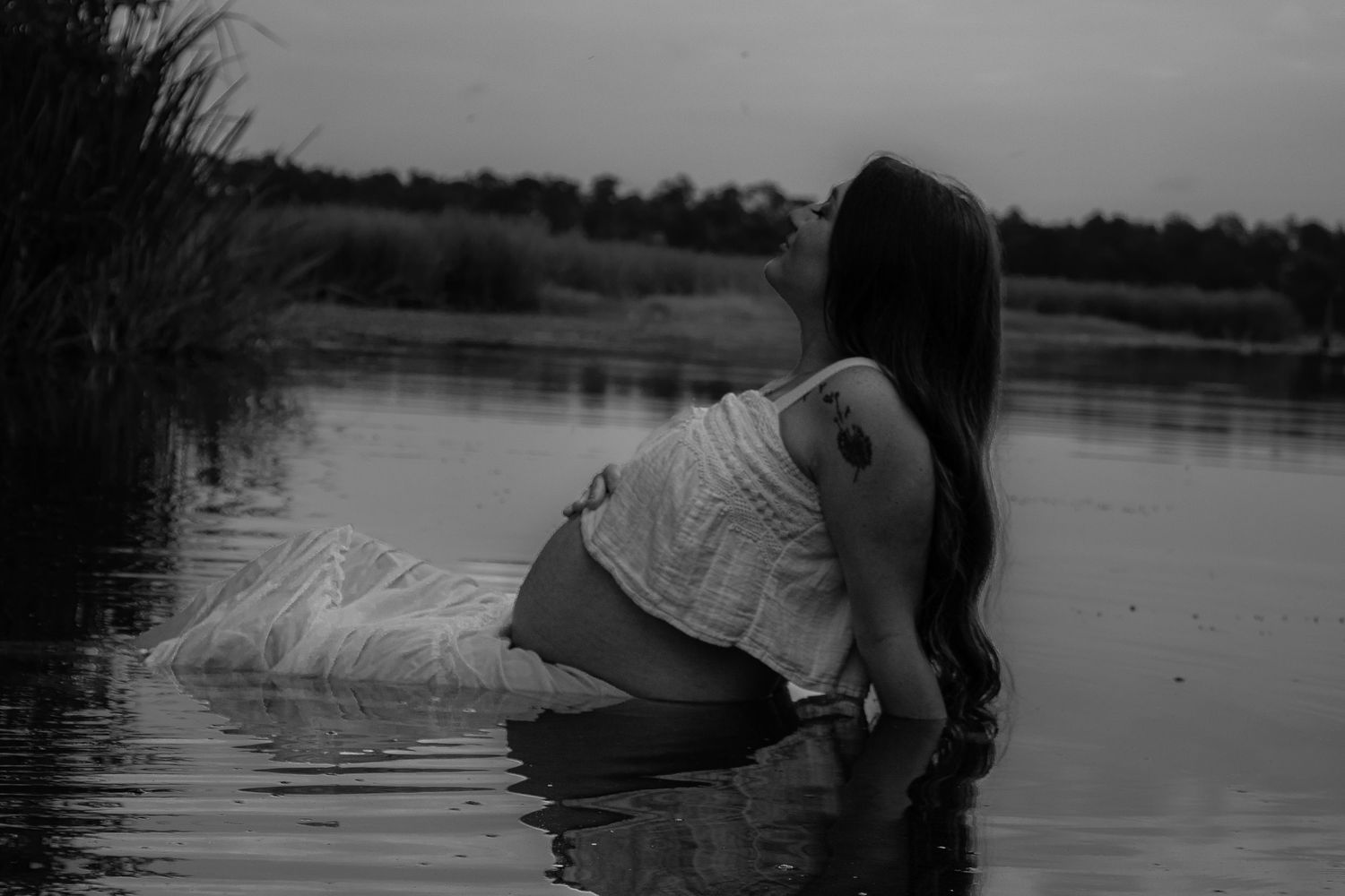 Black and white photo of pregnant woman in white dress sitting in calm lake water at dusk.