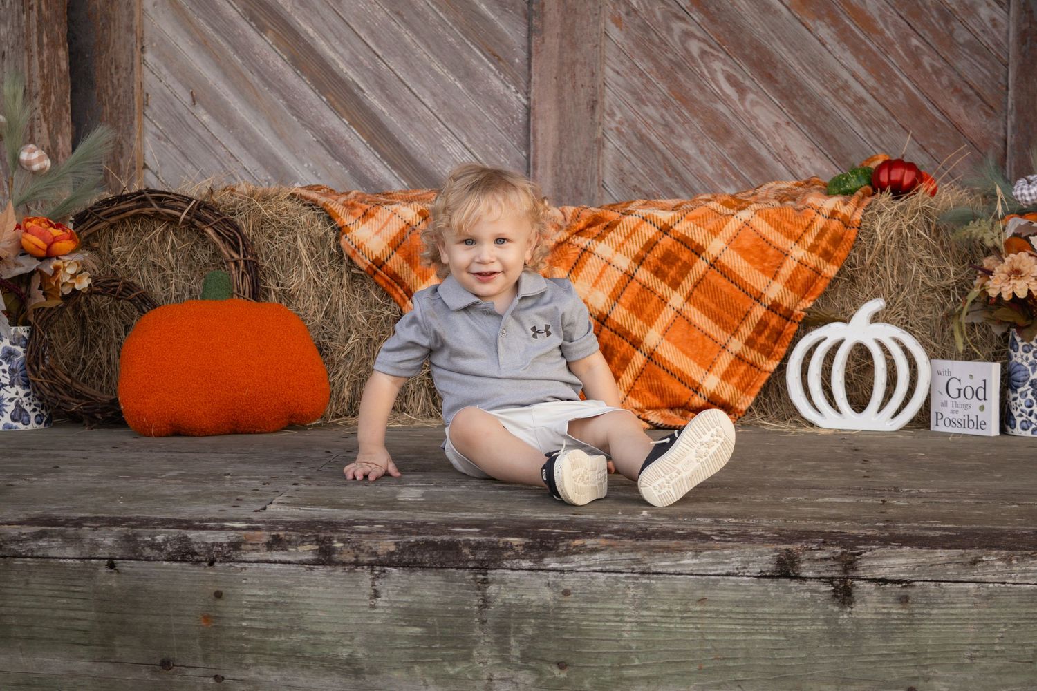 Smiling toddler boy in gray outfit sitting on wooden porch with fall harvest decorations.
