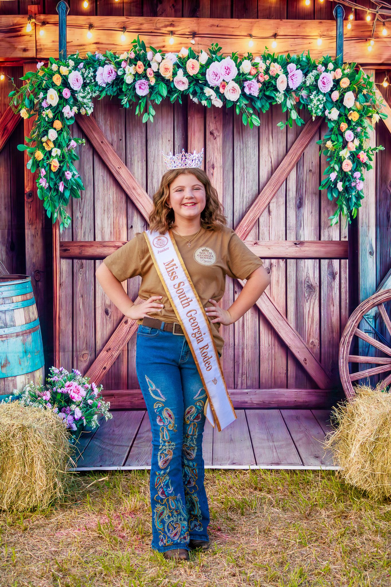Teen beauty queen in crown and sash poses before floral barn door backdrop with hay bales.