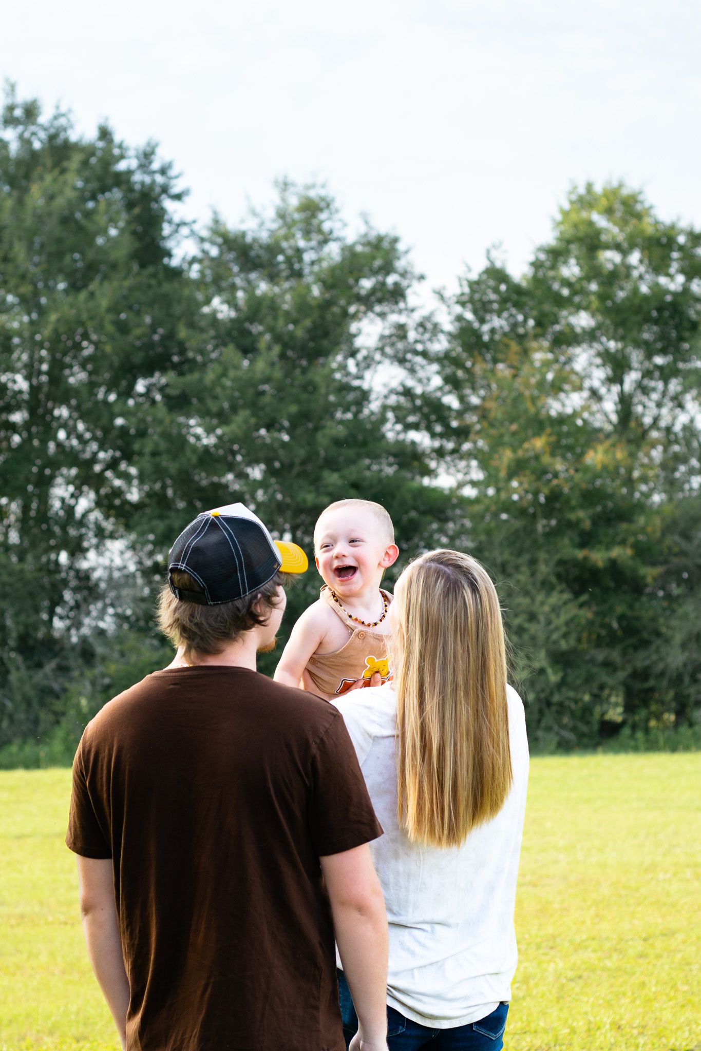 A joyful baby laughing outdoors while being held by parents on a green lawn with trees in the background.