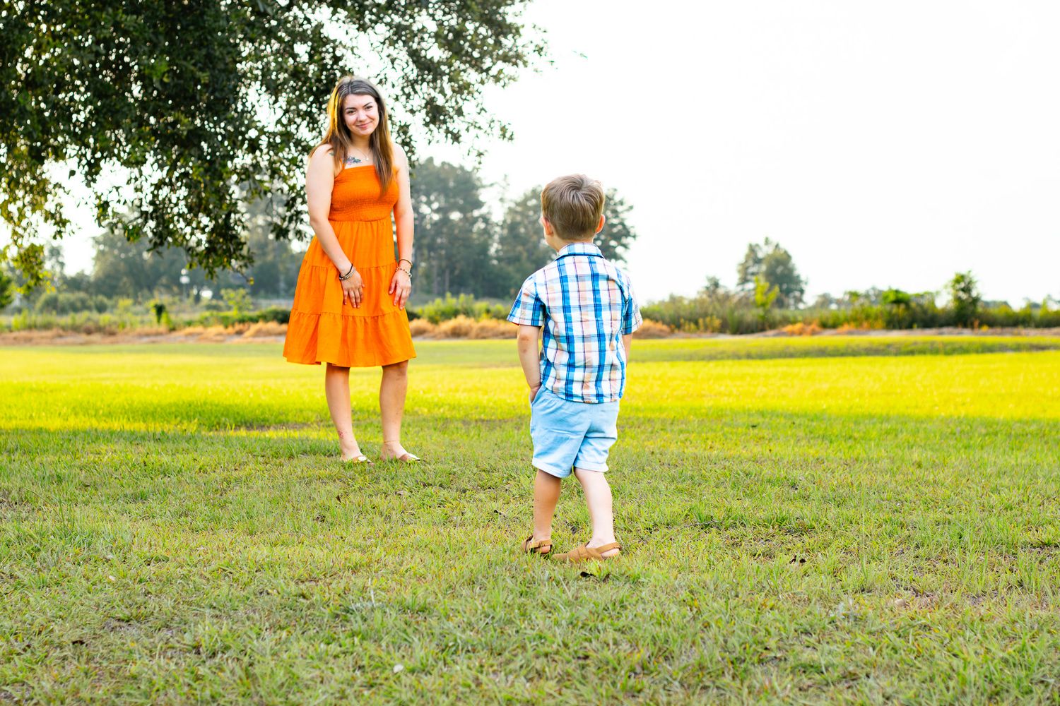 A woman in an orange dress stands barefoot on green grass as a young boy in a plaid shirt runs toward her outdoors.