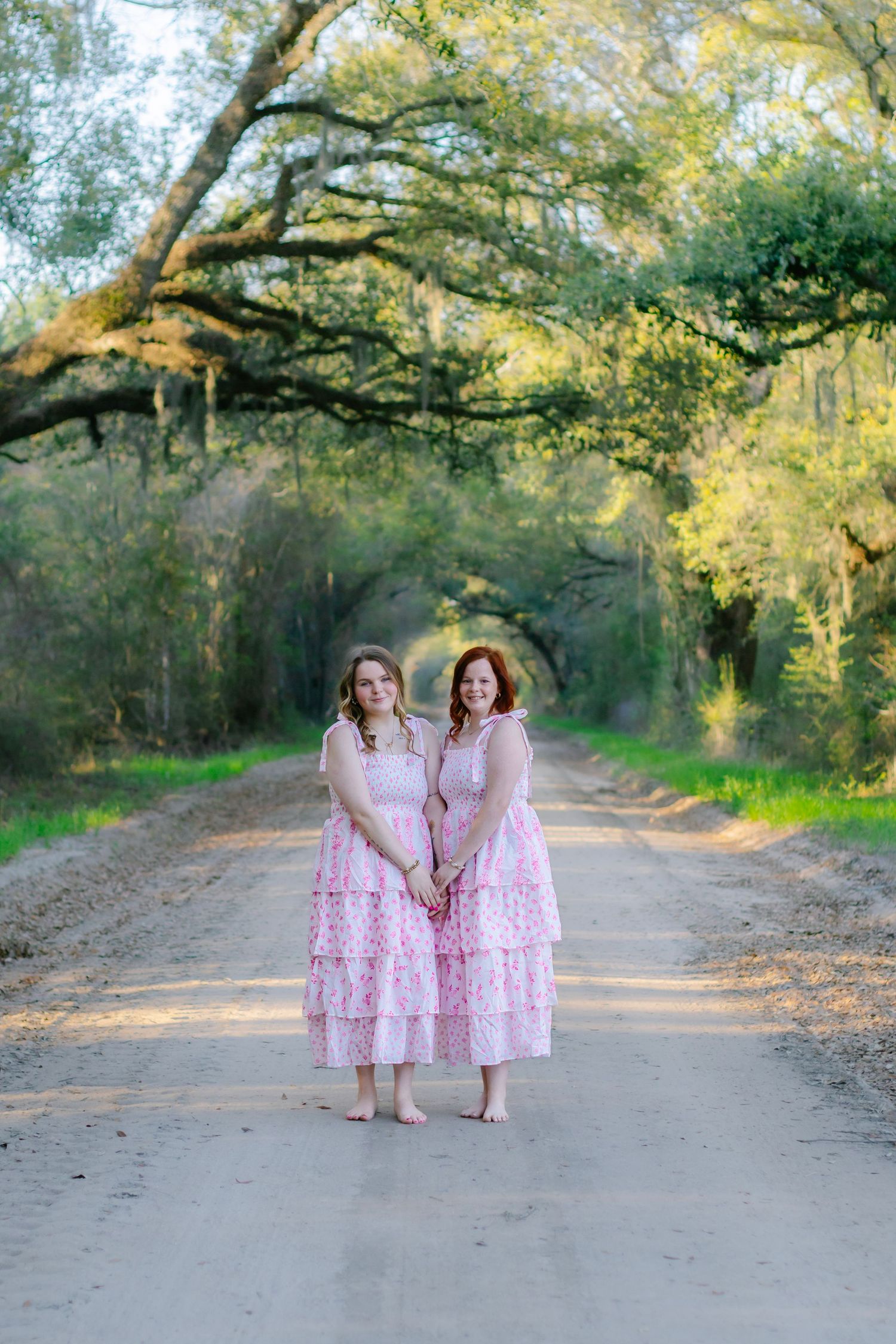 Two young women in matching pink floral dresses stand barefoot on a sunlit tree-lined dirt road.