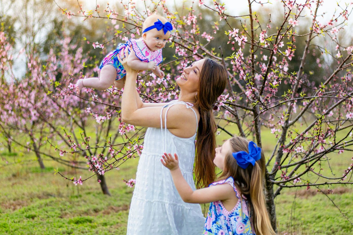Mother lifting baby girl as older daughter watches near pink blossoming trees.