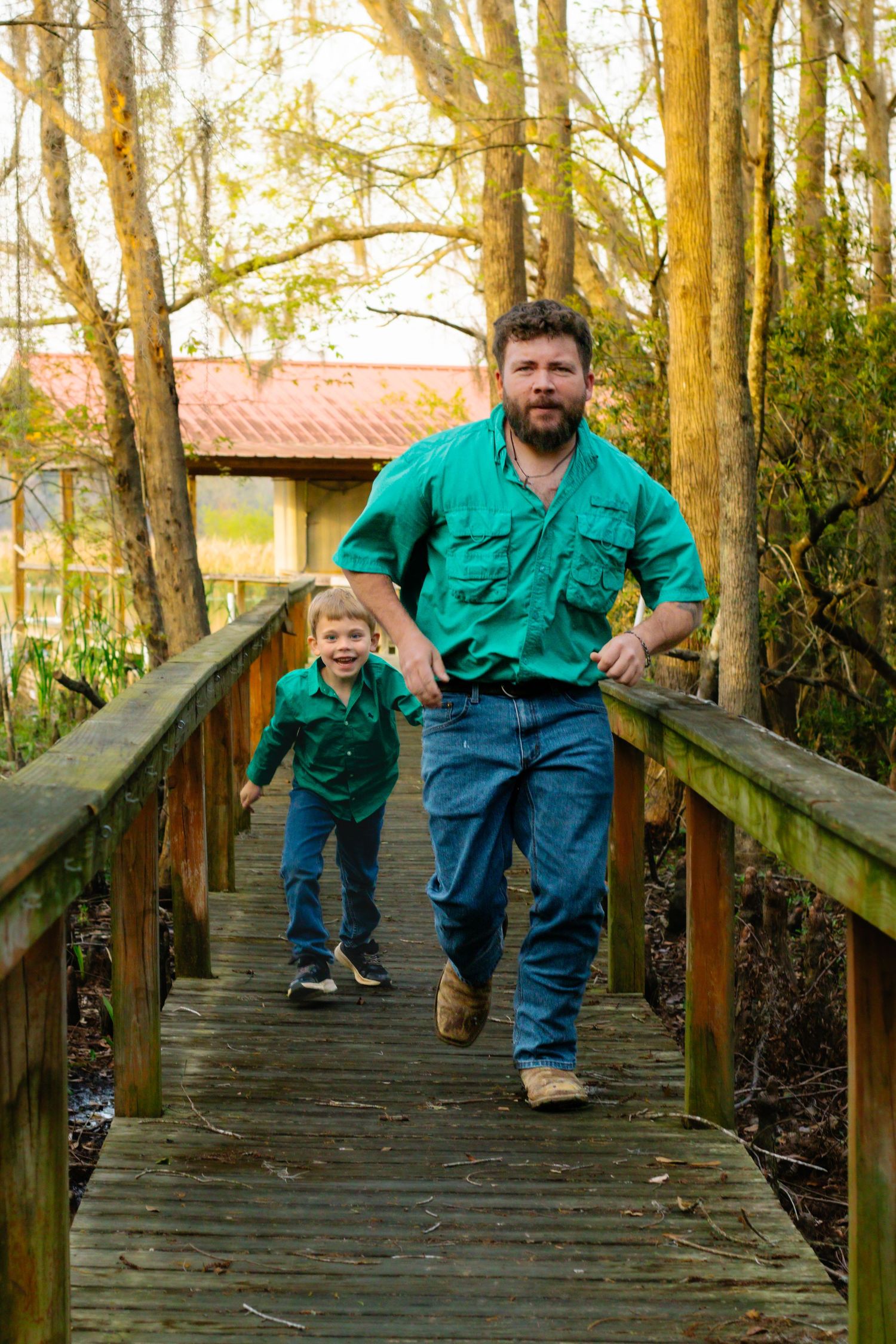 Father and son in matching green shirts running together on a wooden boardwalk outdoors.