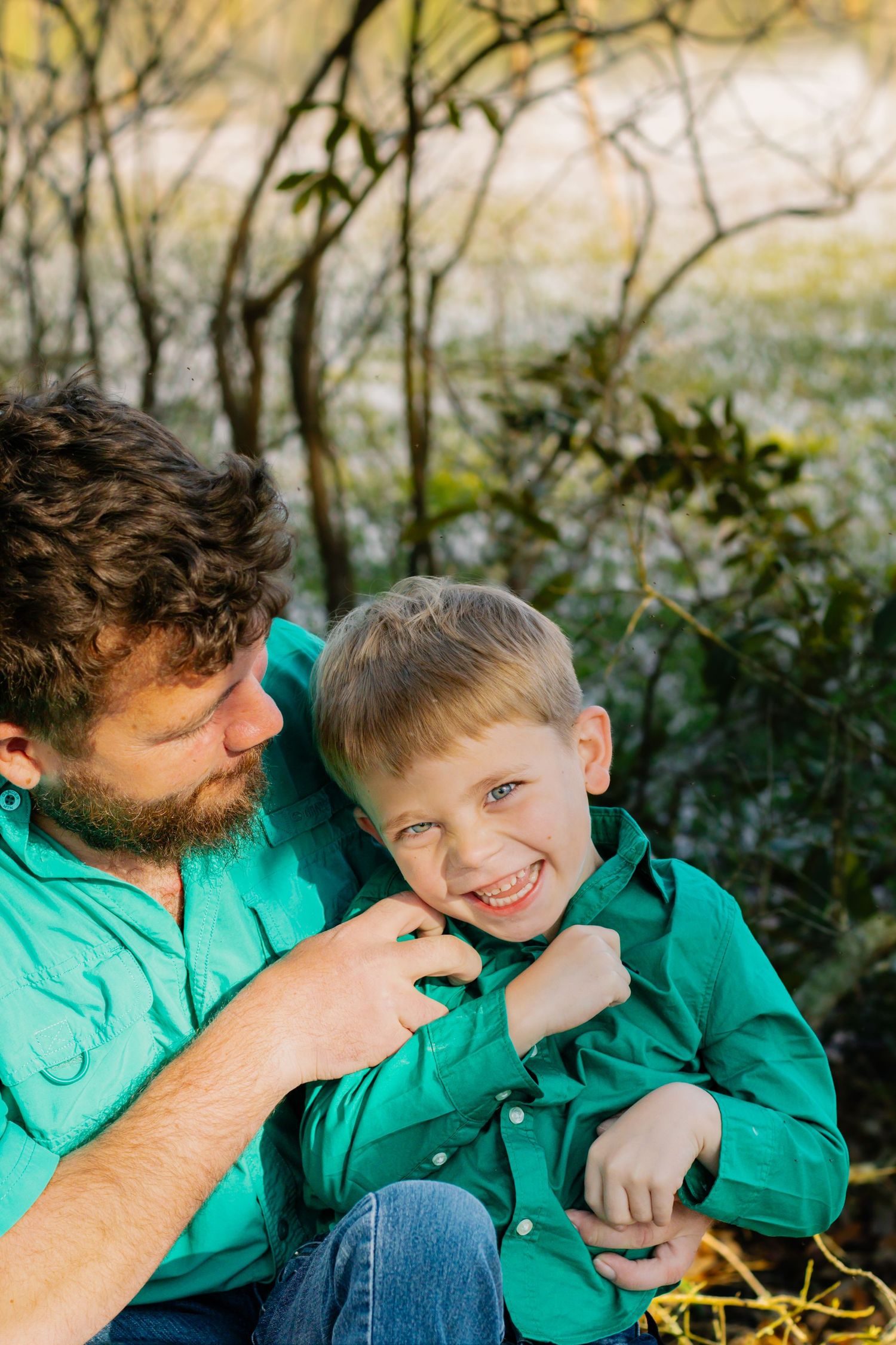 A bearded father in a green shirt playfully interacts with his smiling young son outdoors in nature.