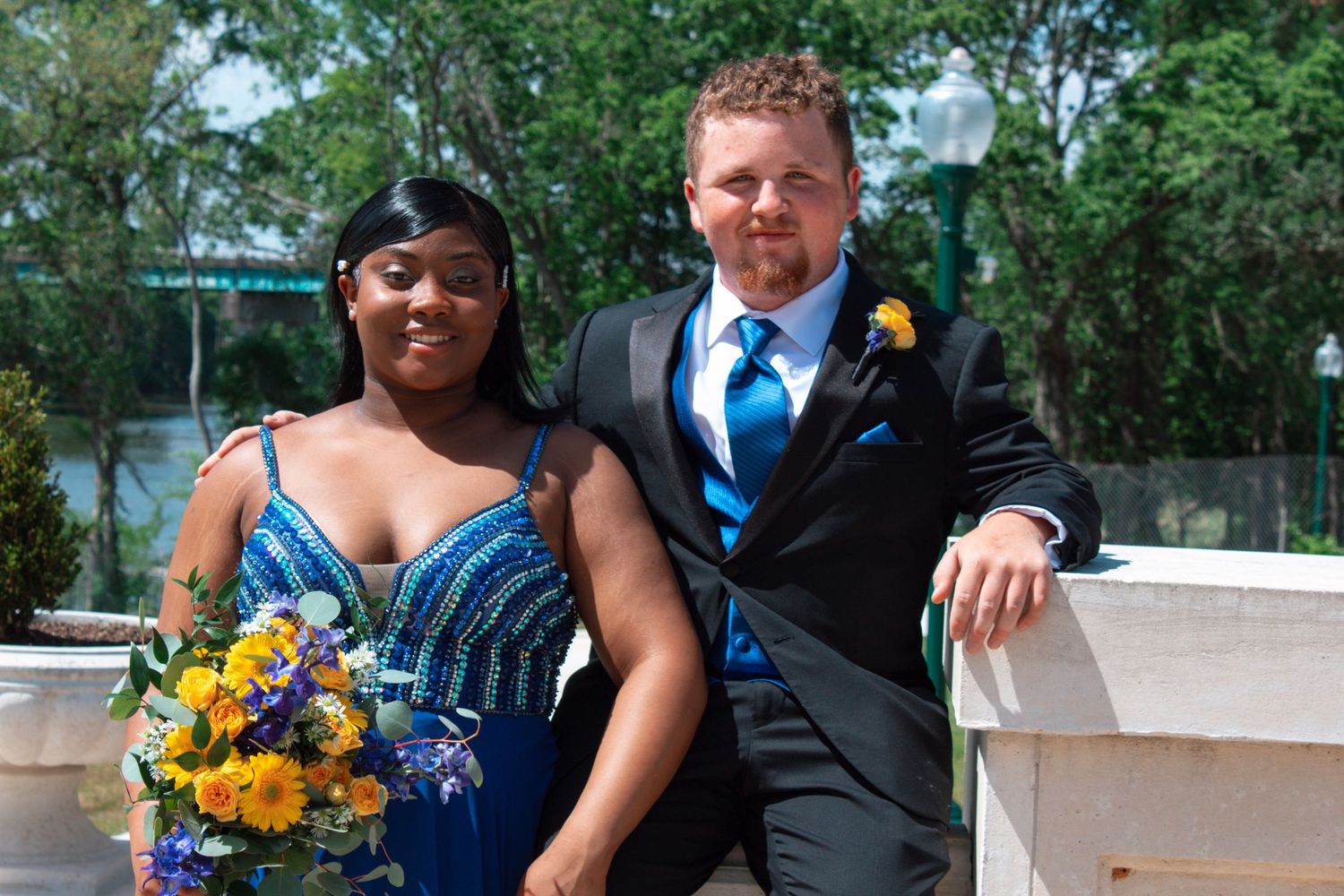A couple in blue prom attire poses outdoors, man in tux with boutonniere, woman holding bouquet.