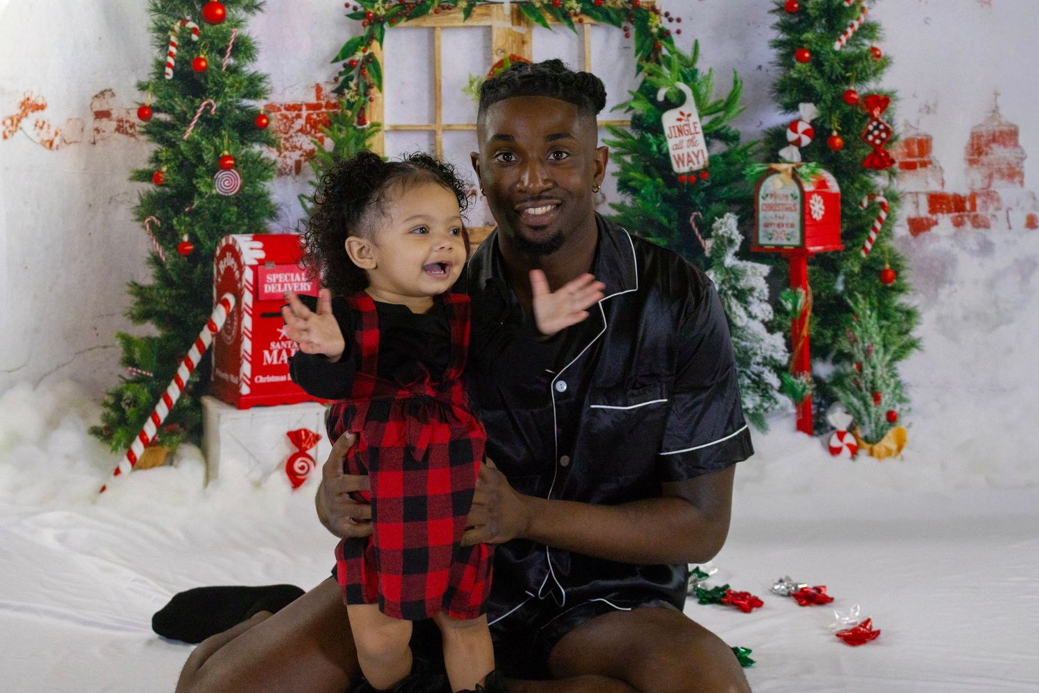 Man in black satin pajamas smiling with a young child in a red plaid dress during a Christmas photo session.