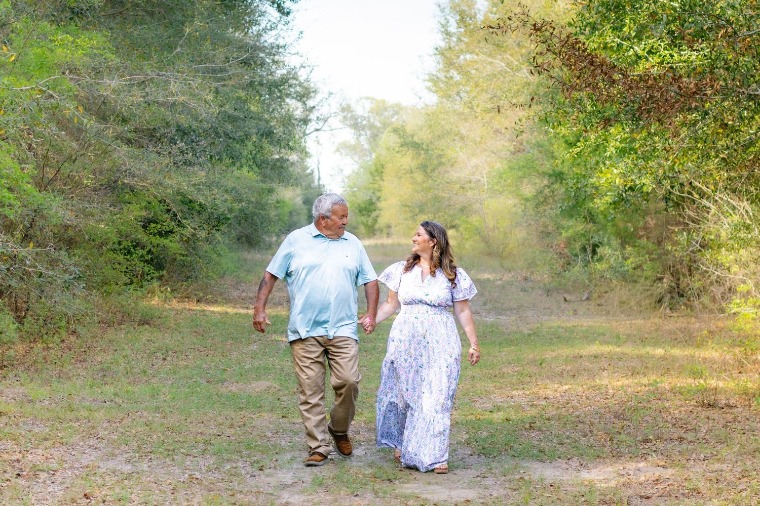 Couple holding hands and walking along a sunny wooded path, woman in floral maxi dress.