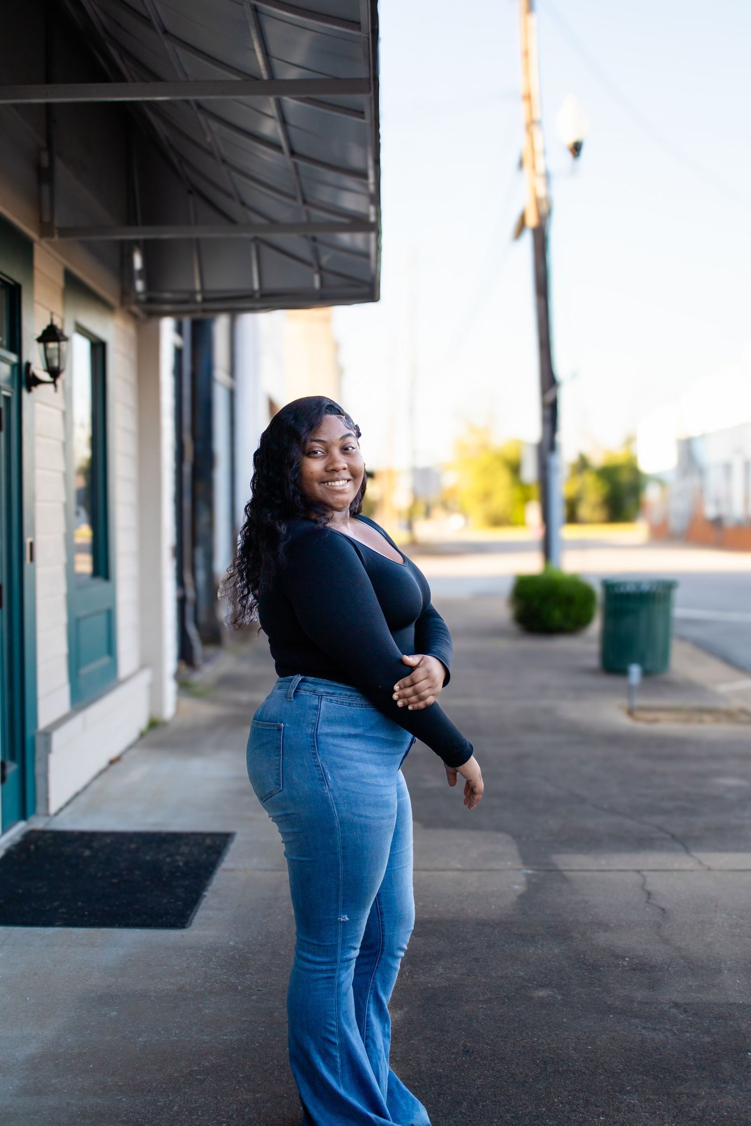 Smiling young Black woman in black top and blue jeans posing on a small town sidewalk.