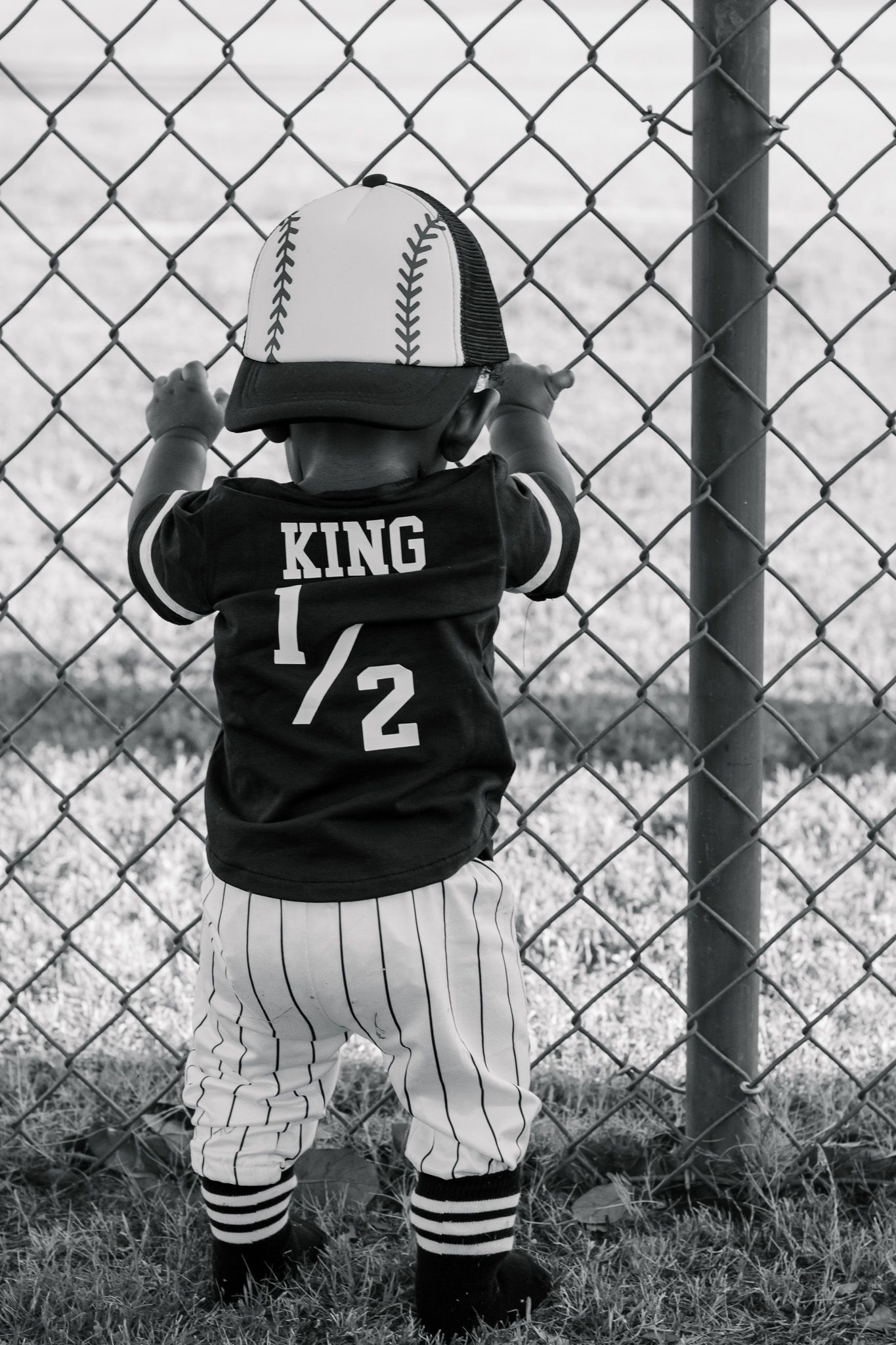 Black and white photo of a toddler in a baseball jersey reading KING 1/2 holding a chain-link fence.