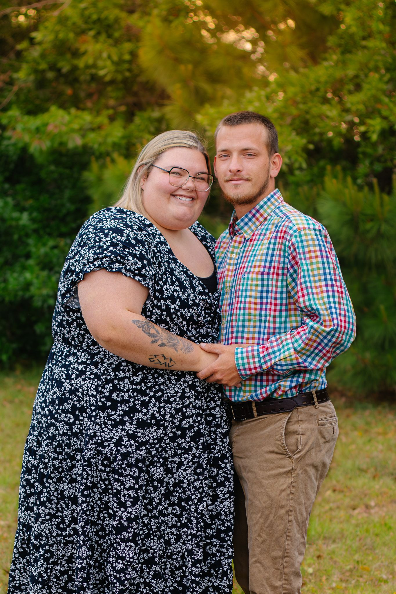 A couple poses outdoors holding hands, woman in floral dress with tattoo, man in colorful plaid shirt, green trees behind them.