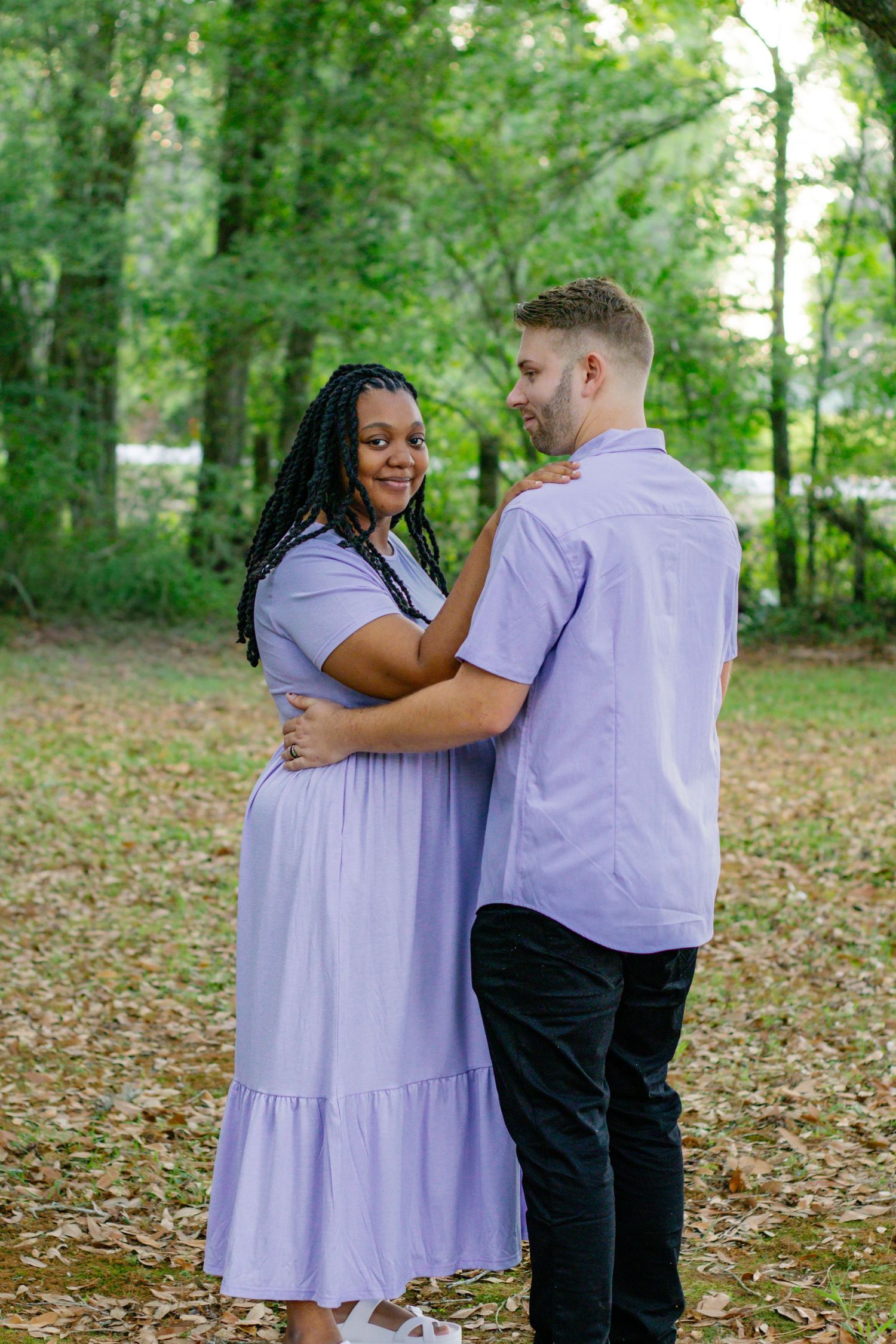 Happy interracial couple in matching purple outfits share a romantic embrace in a wooded outdoor setting.