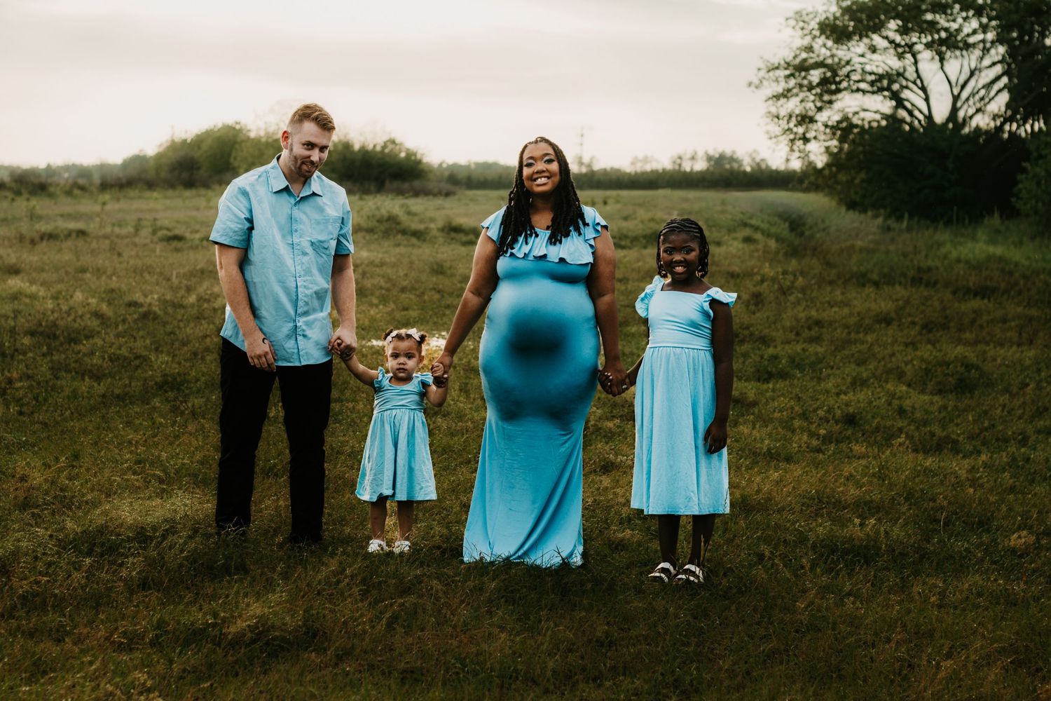 Interracial family of four in coordinating blue outfits holds hands in open green meadow.