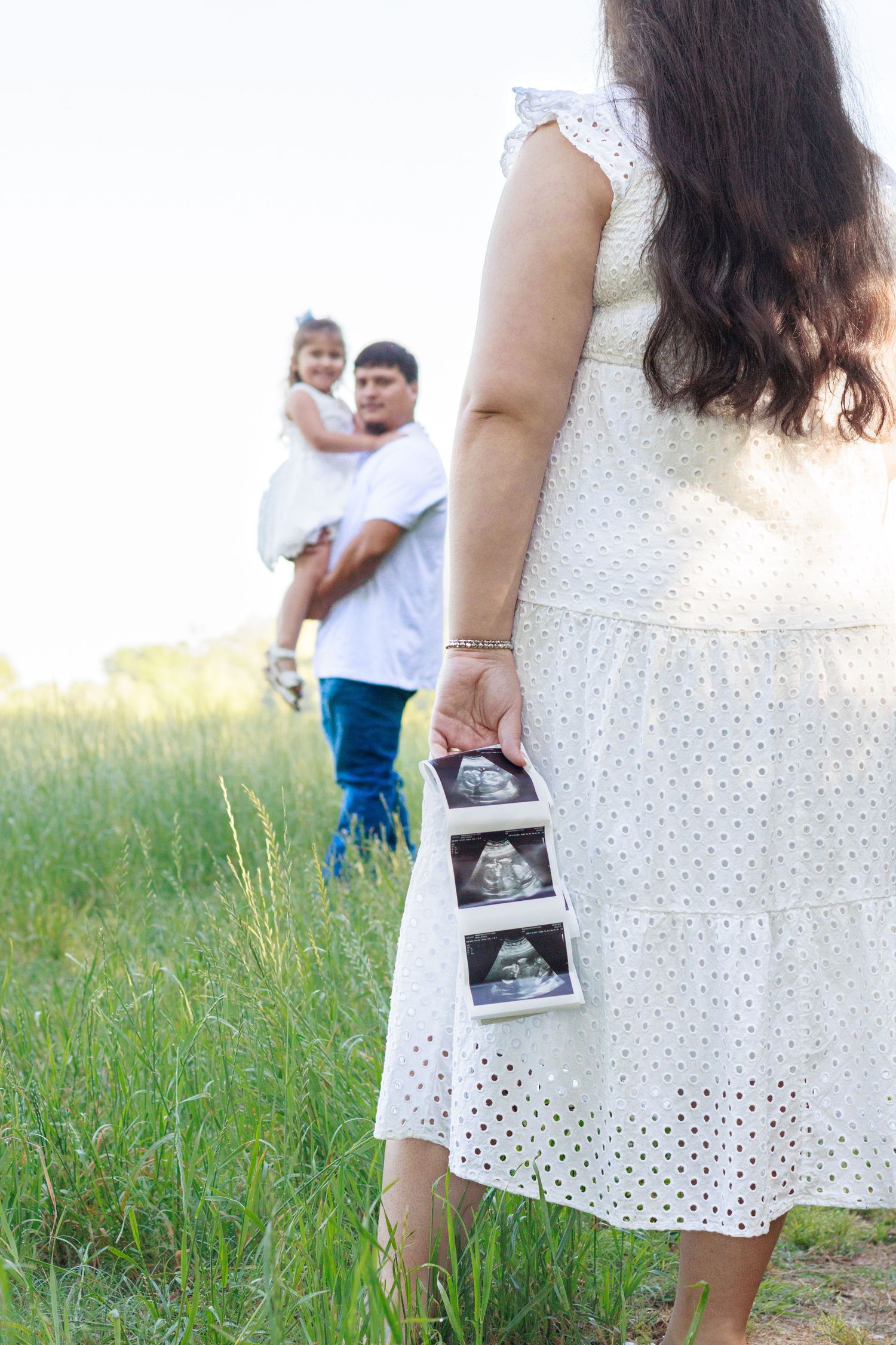 Pregnant woman in white dress holding ultrasound photos in a green field with family in background.