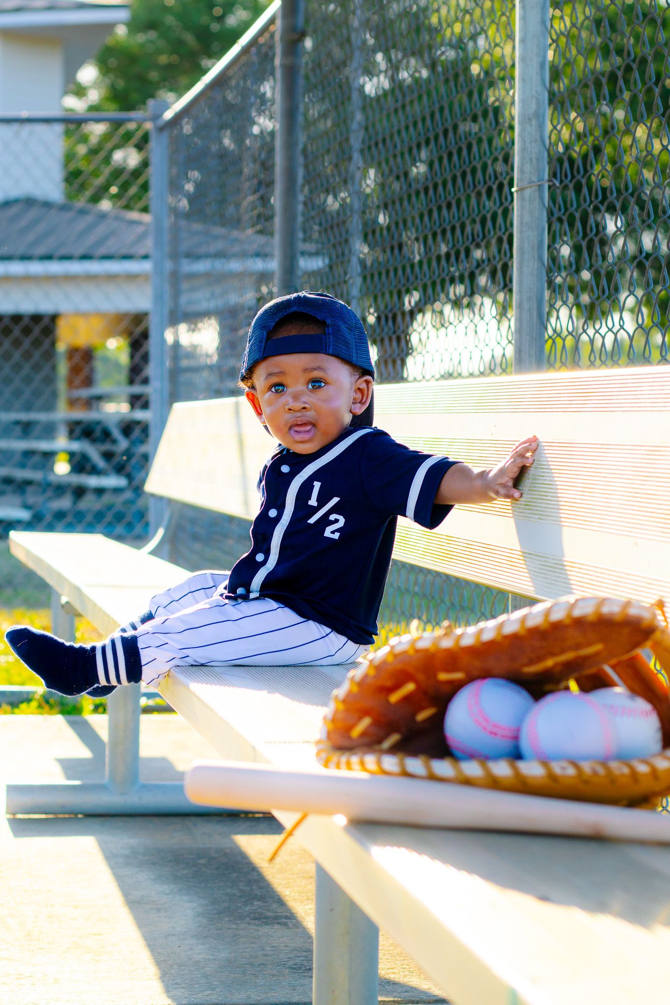 Toddler in baseball uniform sitting on dugout bench with mitt and baseballs.