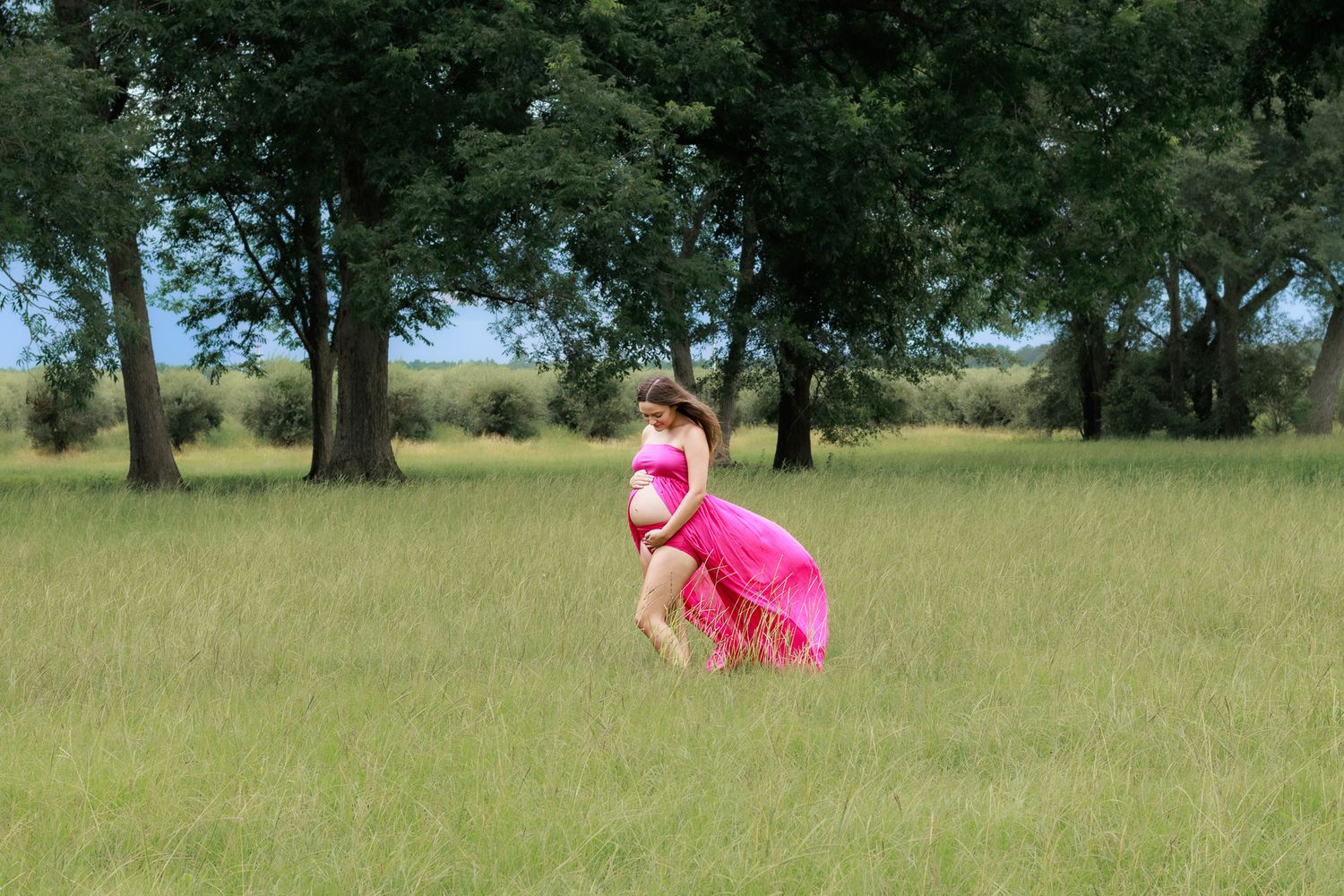 Pregnant woman in flowing pink dress walking through a green meadow with trees in the background.