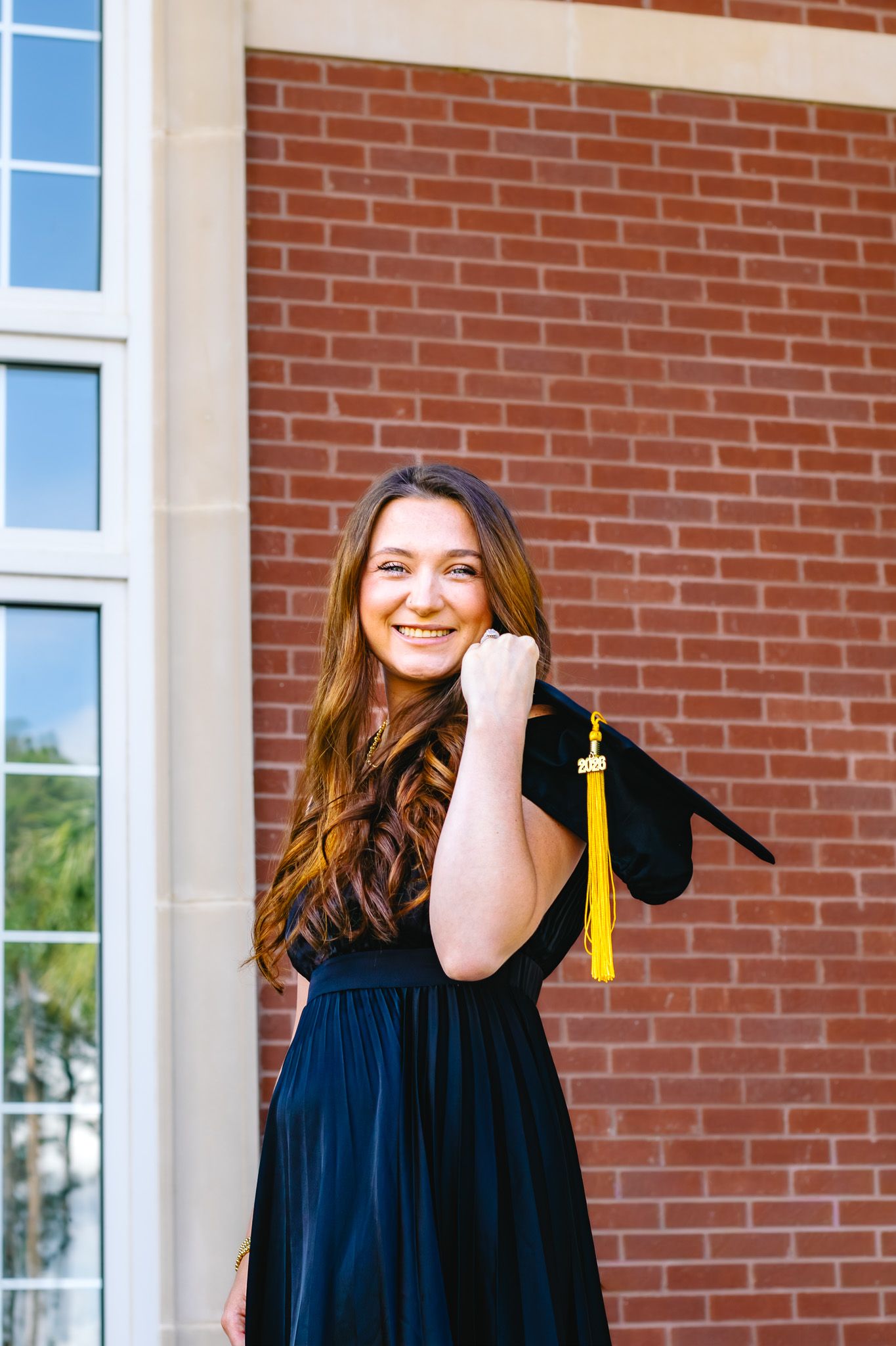 Happy young woman in black graduation gown holds cap with gold tassel against brick wall outdoors.