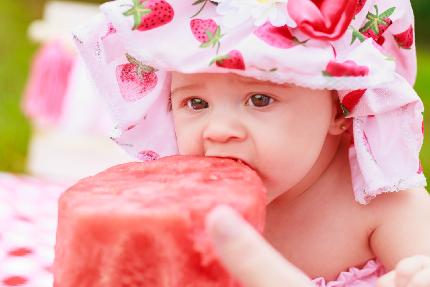 Adorable baby in a strawberry sun hat eagerly eating a fresh slice of watermelon outdoors in summer.