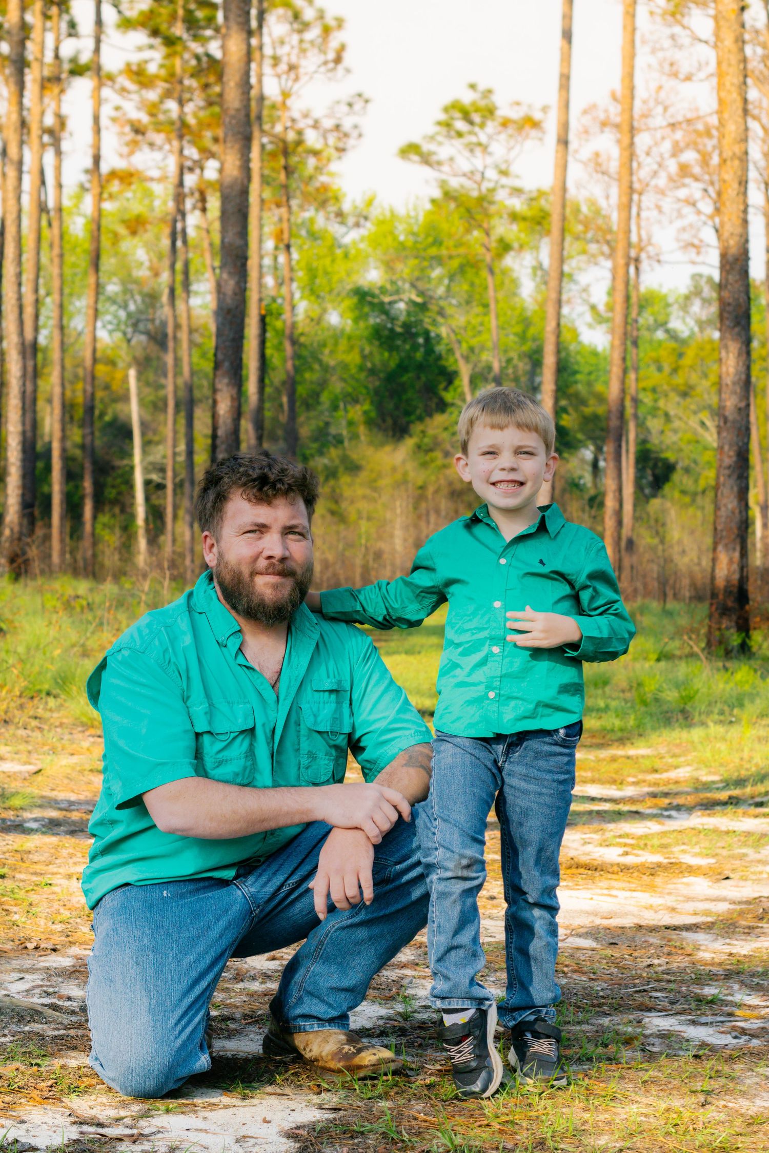 A bearded father kneels beside his smiling young son on a sunny forest path, both wearing green shirts.