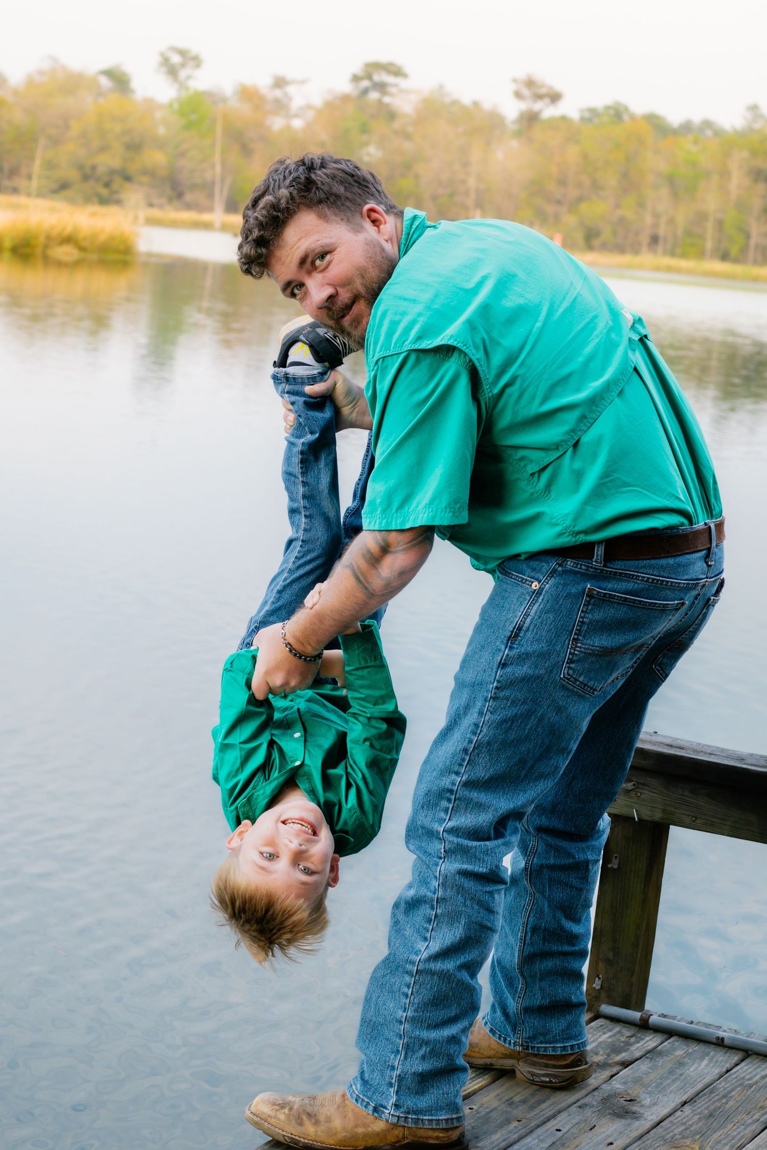 Laughing father playfully holds young son upside down on a wooden dock by a lake.