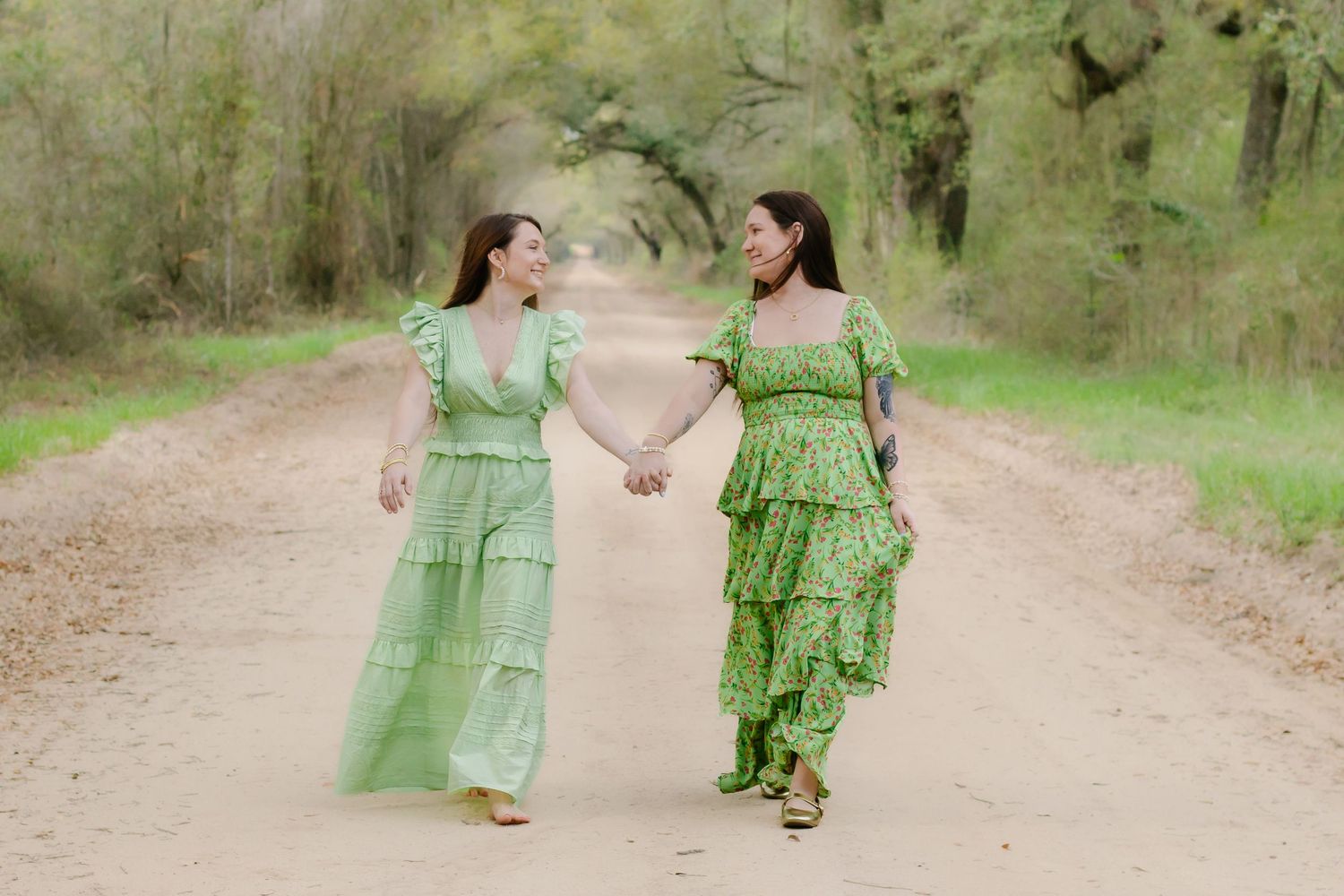 Two women in matching green dresses holding hands and walking down a scenic tree-lined dirt path.