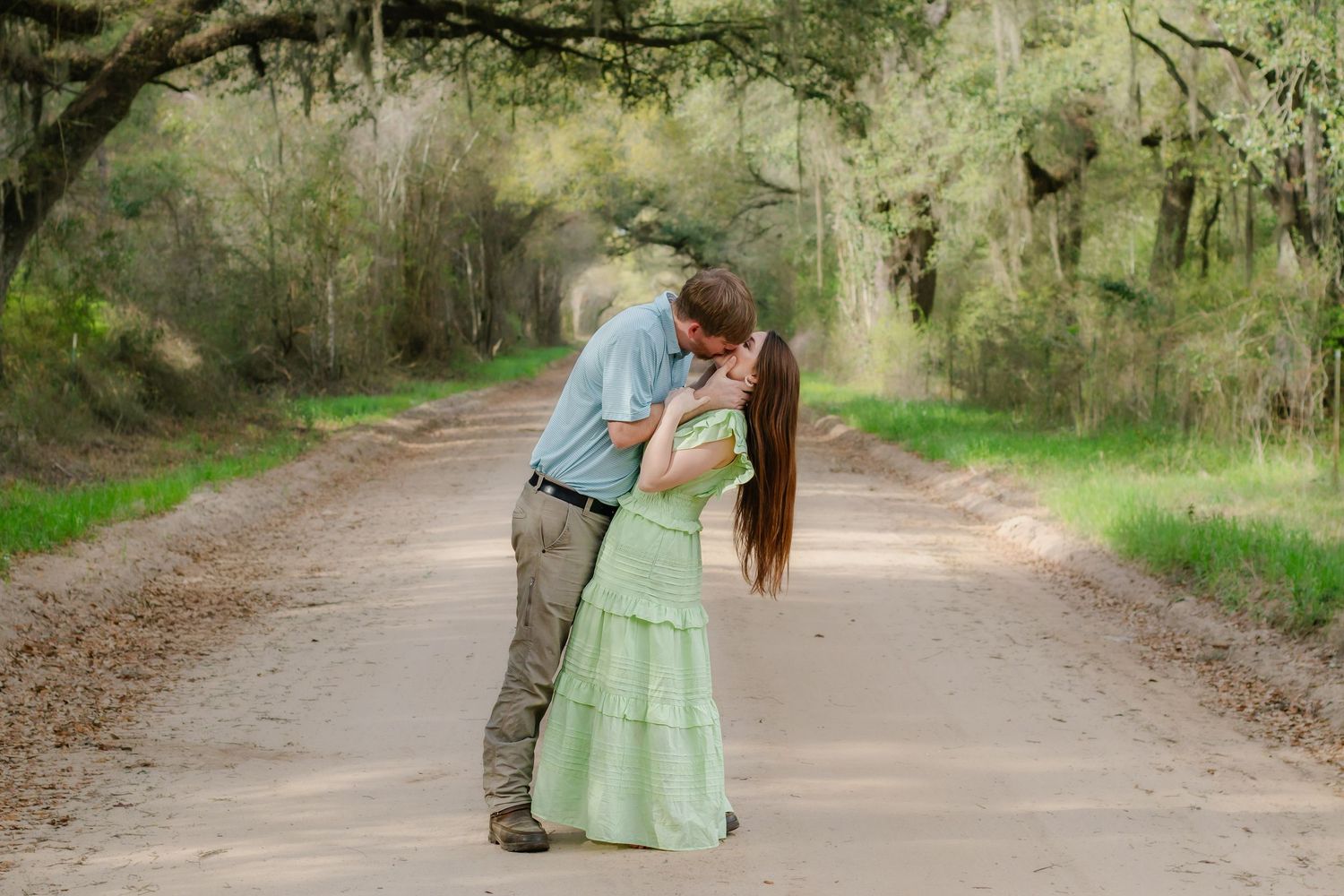 Couple sharing a tender kiss on a scenic dirt road canopied by large oak trees with Spanish moss.