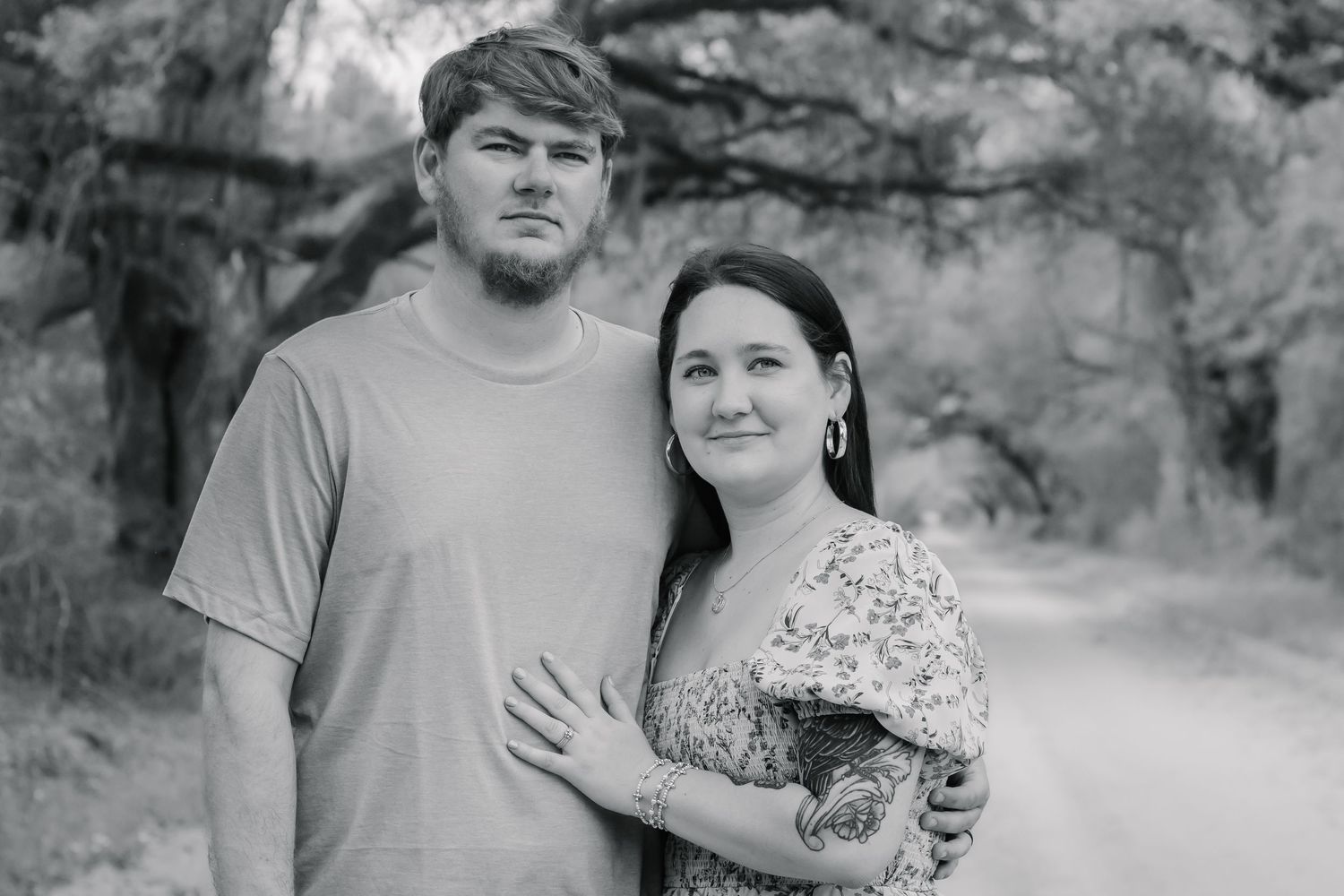 Black and white photo of a couple embracing on a wooded dirt road, woman in floral dress leaning against mans shoulder.