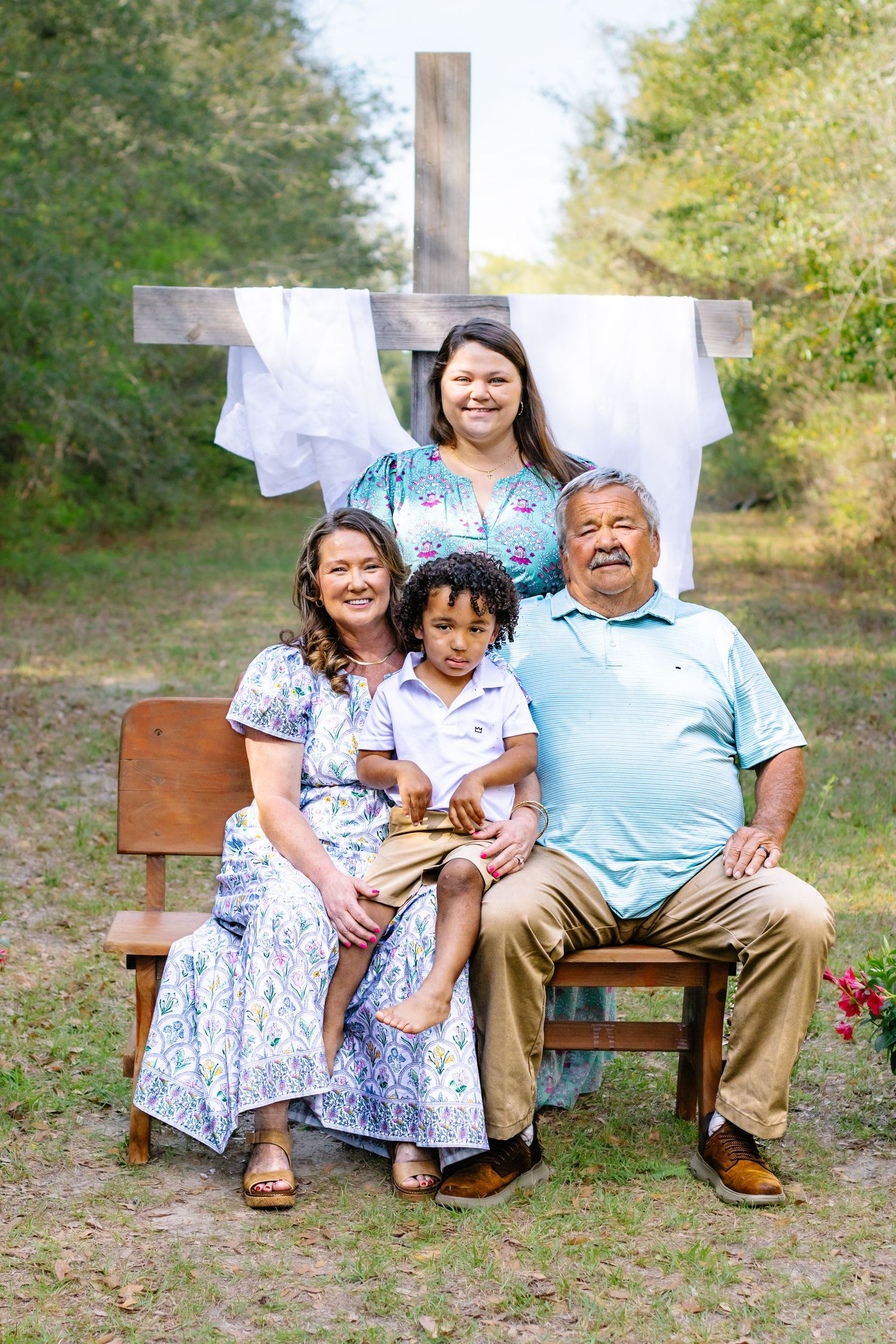 Family of four with young boy sitting on bench in front of a rustic wooden cross in outdoor setting.