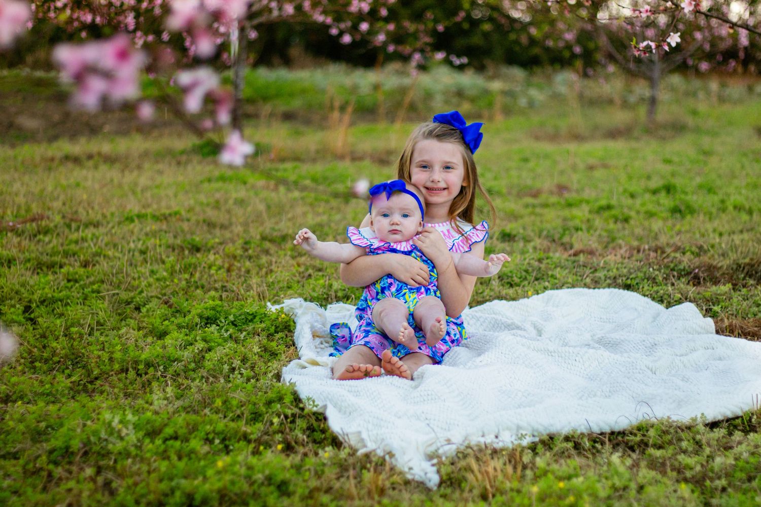 Two sisters sitting on white blanket in grass near pink blossoming trees.