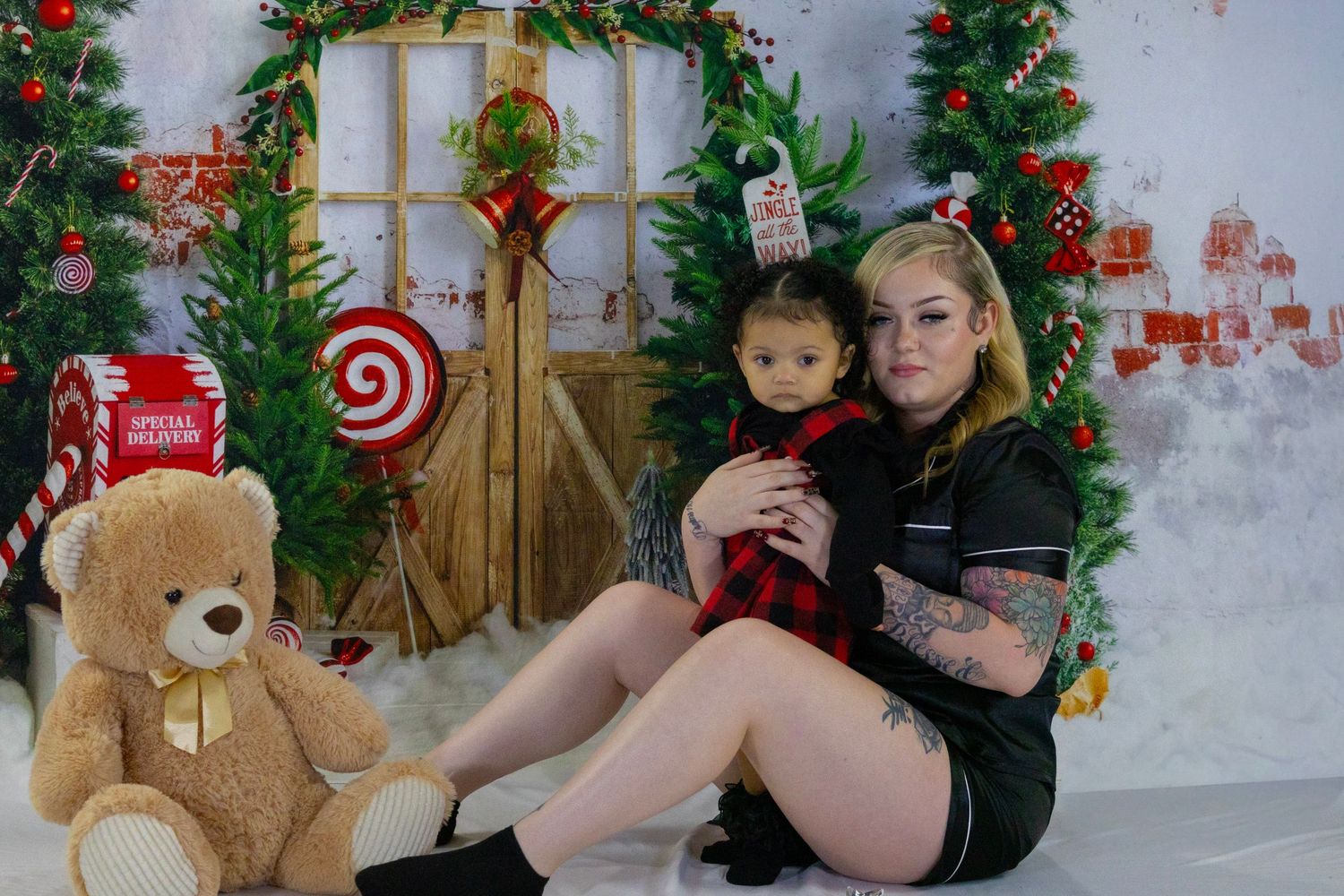 Woman holding a baby in red plaid outfit sitting beside a large teddy bear in a Christmas themed photo setting.
