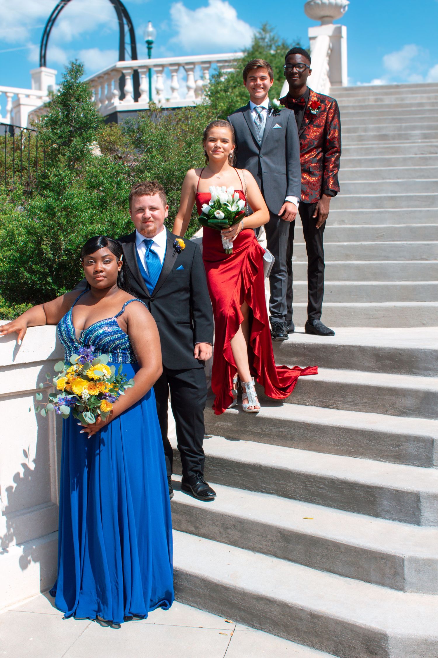 Four prom couples pose on elegant white stairs, dressed in formal attire with bouquets for a special occasion.
