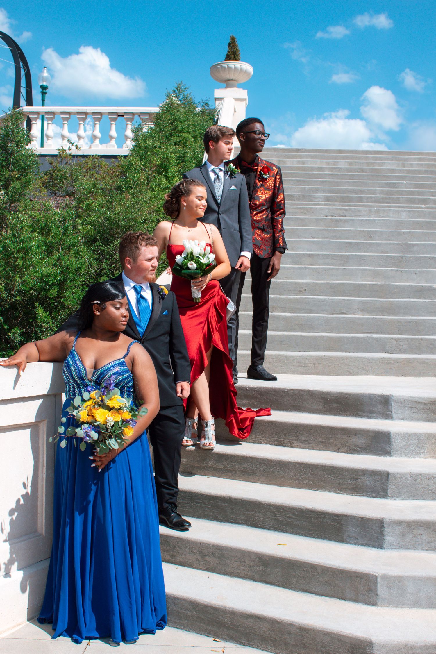 Four prom-goers in formal wear pose on white outdoor stairs under a bright blue sky.