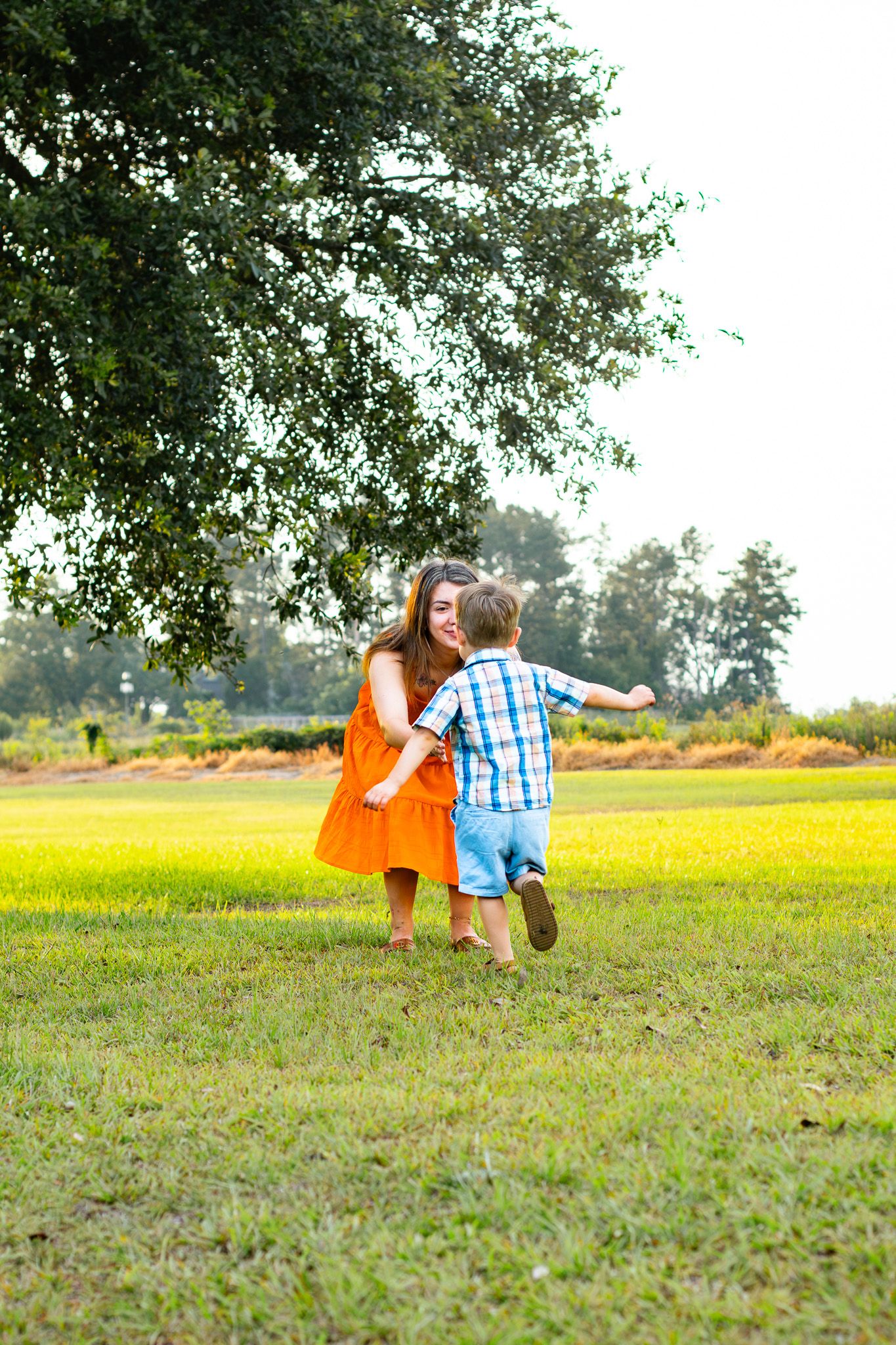 A woman in an orange dress catches a running young boy in a plaid shirt under a large tree outdoors.